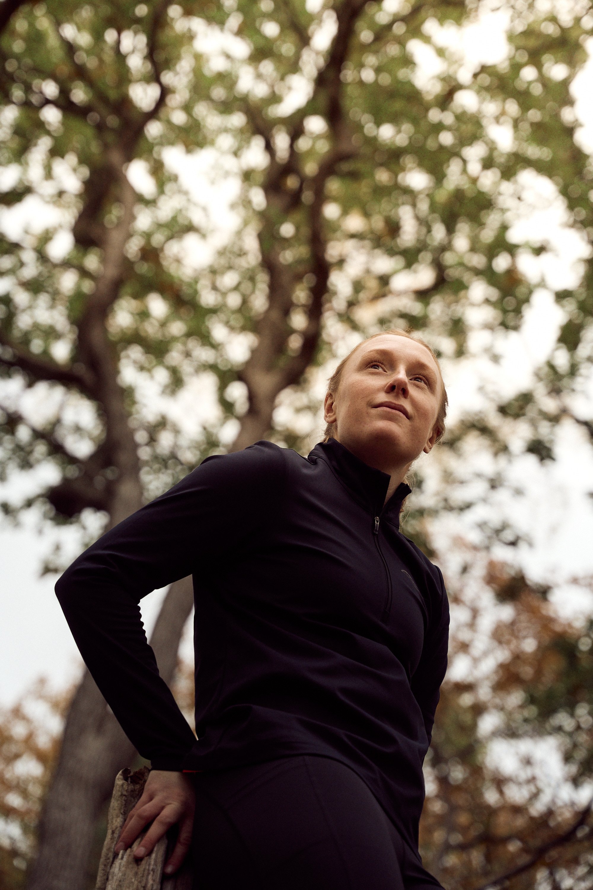 A woman in black athletic wear standing outdoors under a large tree with a cloudy sky background, looking upward, with her hand resting on a piece of wood.