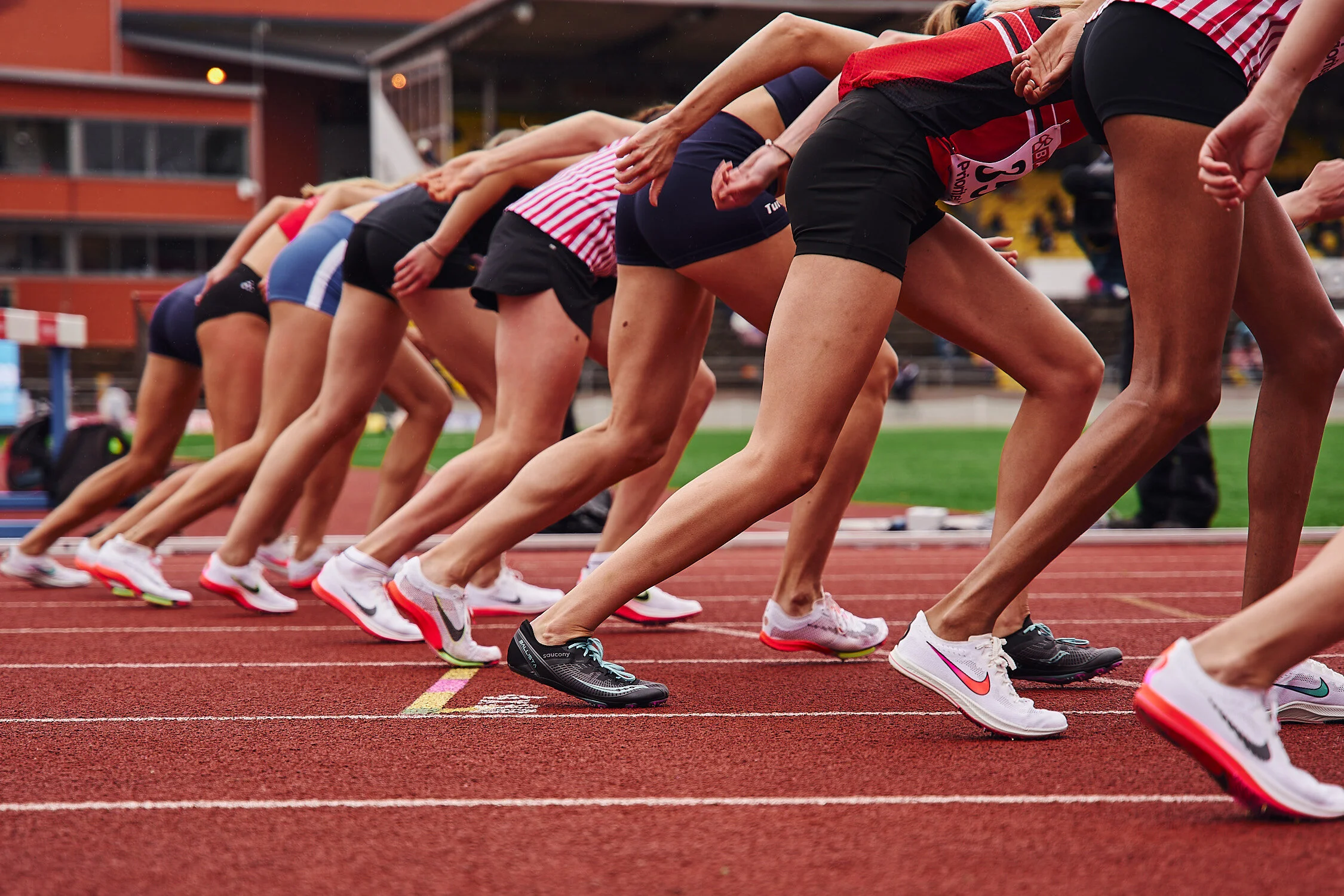 Female athletes in running shoes lined up at the starting line of a track field, preparing for a race.