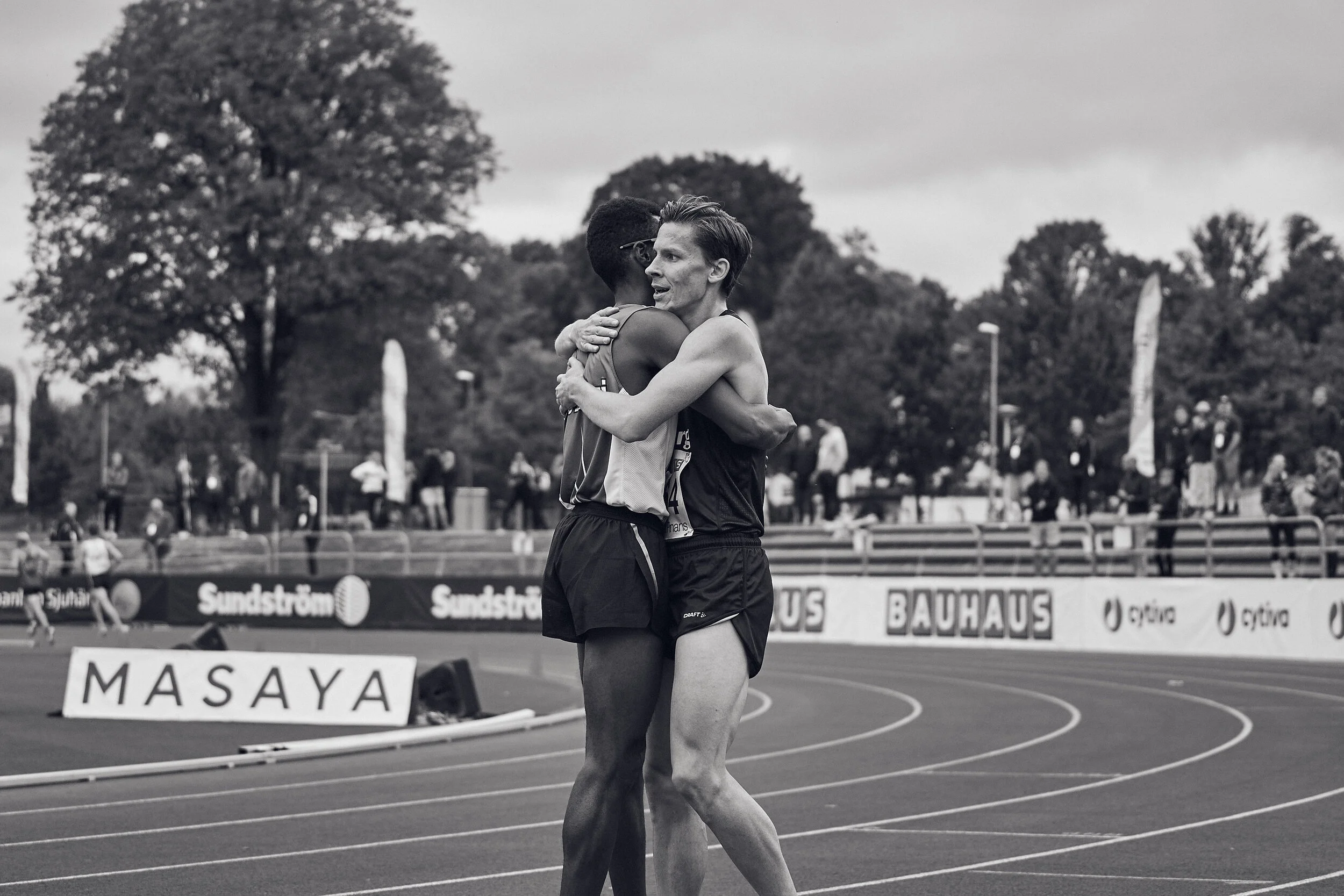 Two male athletes hugging on a running track at an outdoor sports event. One athlete faces the camera with a pleased expression, while the other has his back to the camera. In the background, other athletes and spectators are visible, along with tree