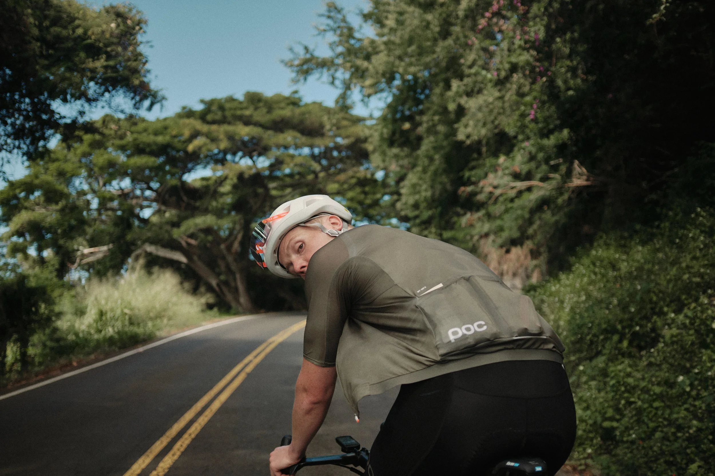 A cyclist wearing a helmet and dark athletic clothing riding on a winding road surrounded by trees.