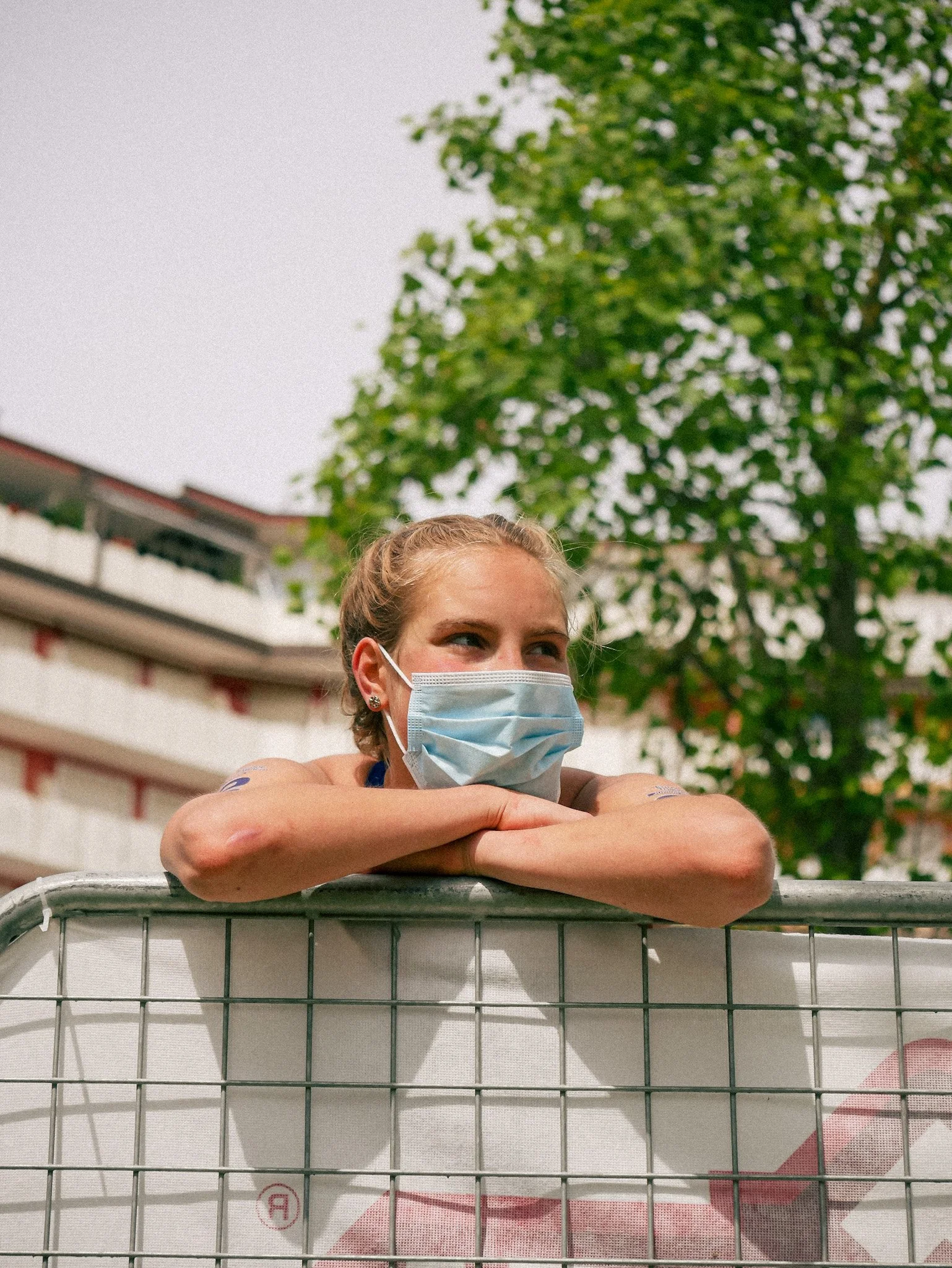 A woman wearing a face mask leans on a white barrier, with her arms crossed and resting on top of it, outdoors with green trees and a building in the background.
