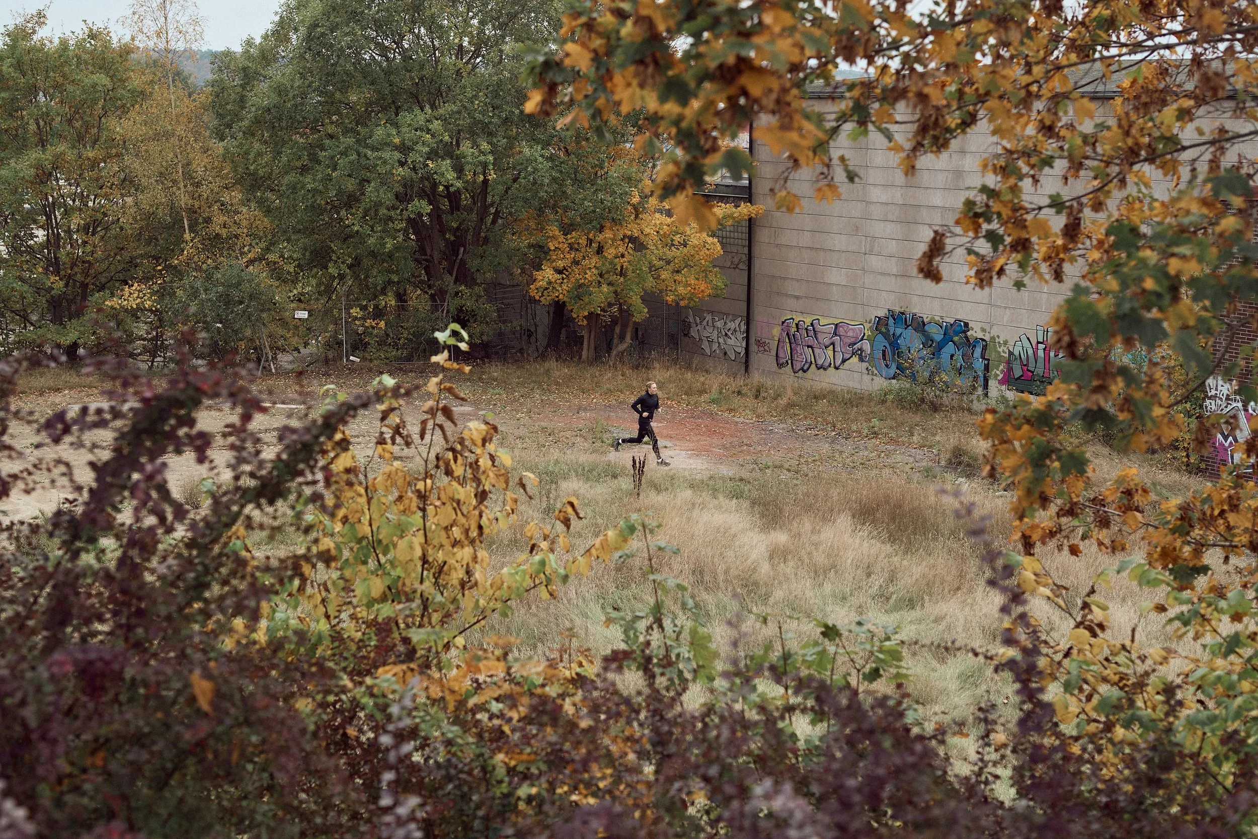 A person jogging in a park with autumn-colored trees and graffiti-covered building wall in the background.