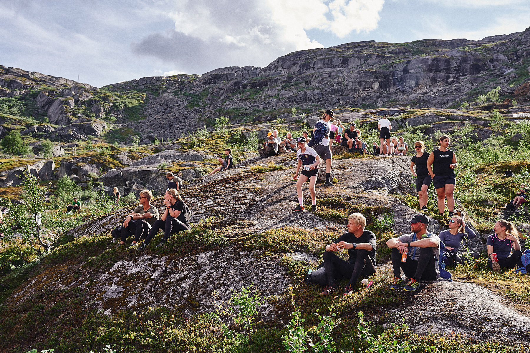 People resting and sitting on a rocky hillside in a mountain landscape with sparse trees and shrubs, under a partly cloudy sky.