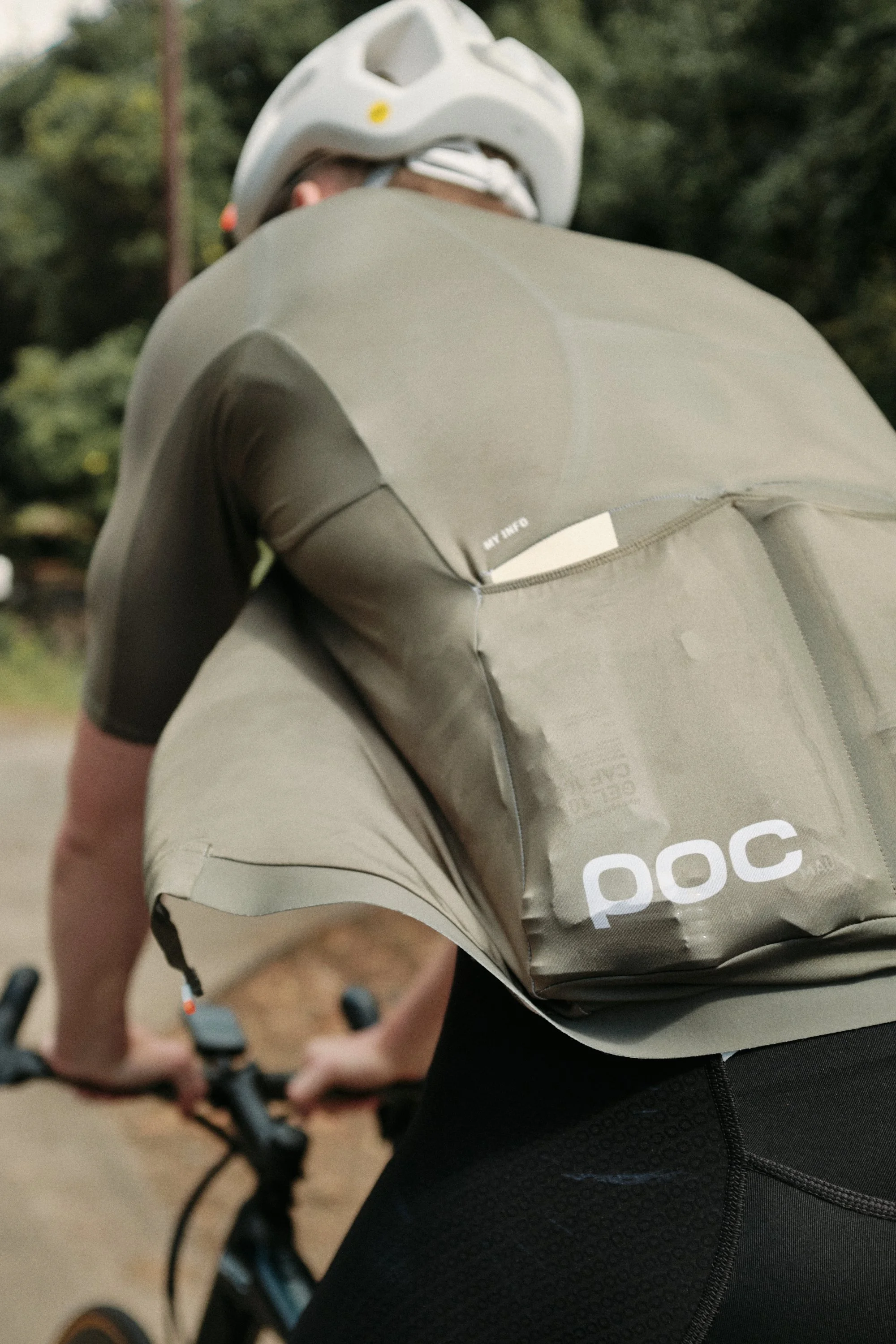 A cyclist wearing a white helmet and a grey cycling jersey, holding the handlebars of a bike outdoors with trees in the background.