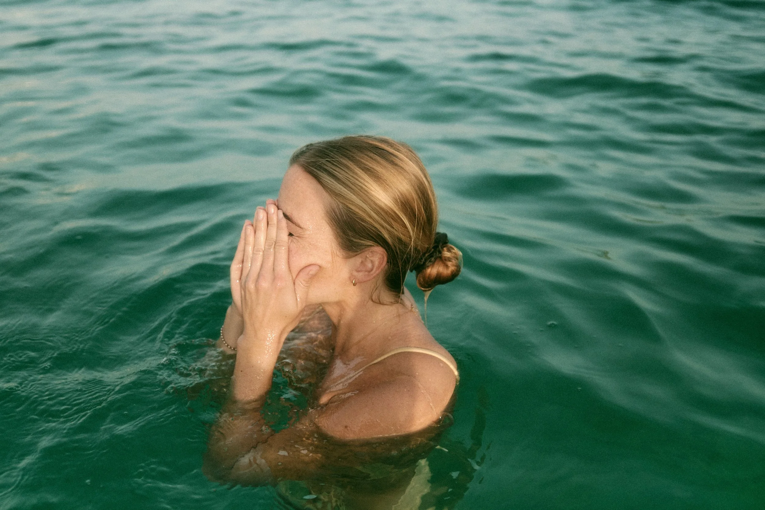 A woman with blonde hair tied in a bun, wearing a beige top, is in the water covering her face with her hand. The water appears to be a greenish ocean or lake, and she is holding her head with both hands.