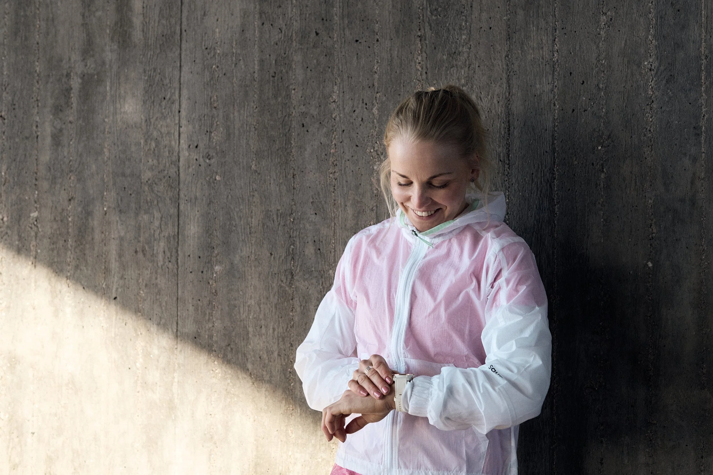A woman standing against a dark textured wall, smiling and looking at her wristwatch