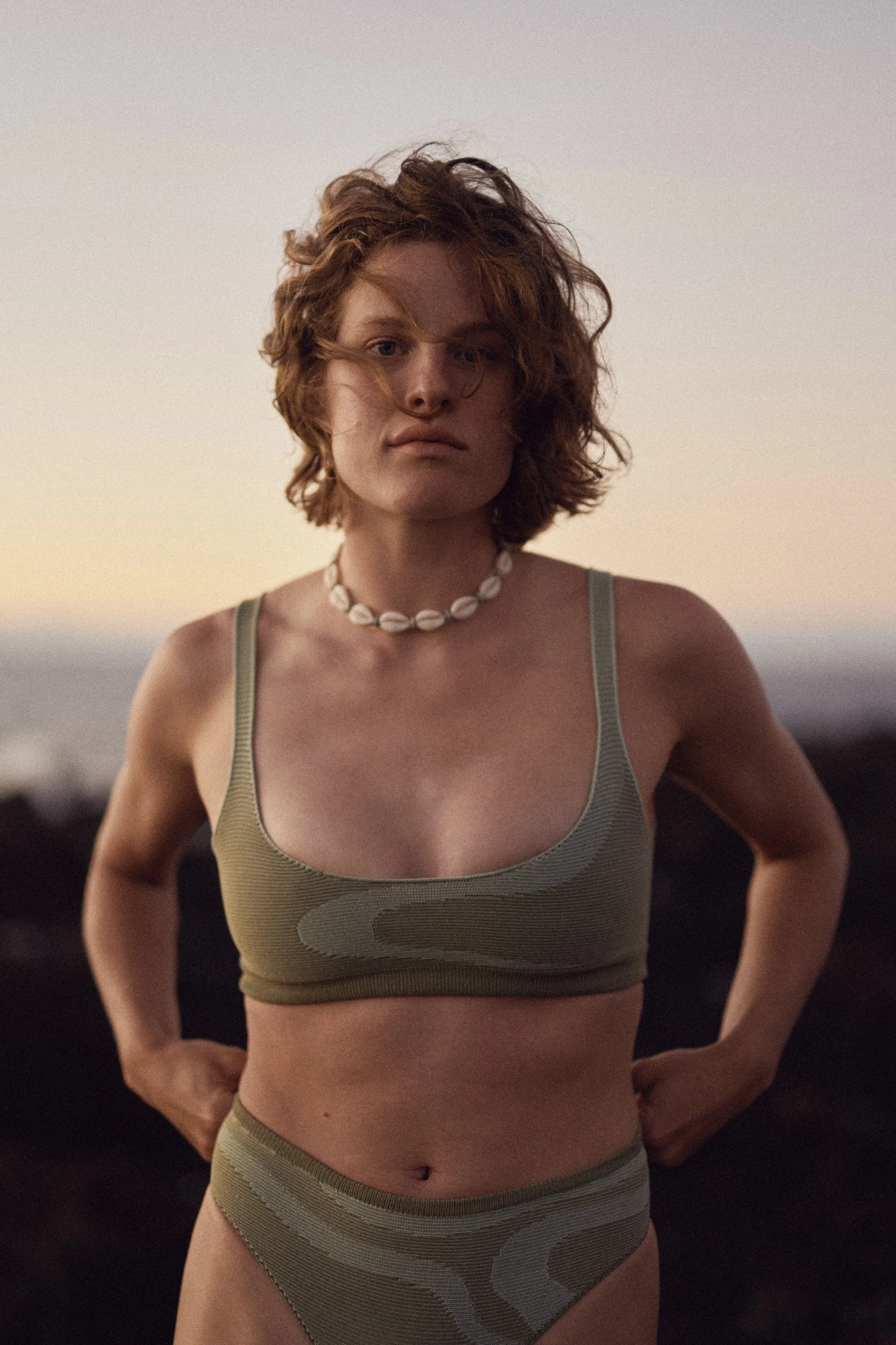 A woman with curly red hair, wearing a green sports bra and matching high-waisted bottoms with a swirl pattern, stands outdoors during sunset with a neutral expression, hands on hips, against a blurred natural background.