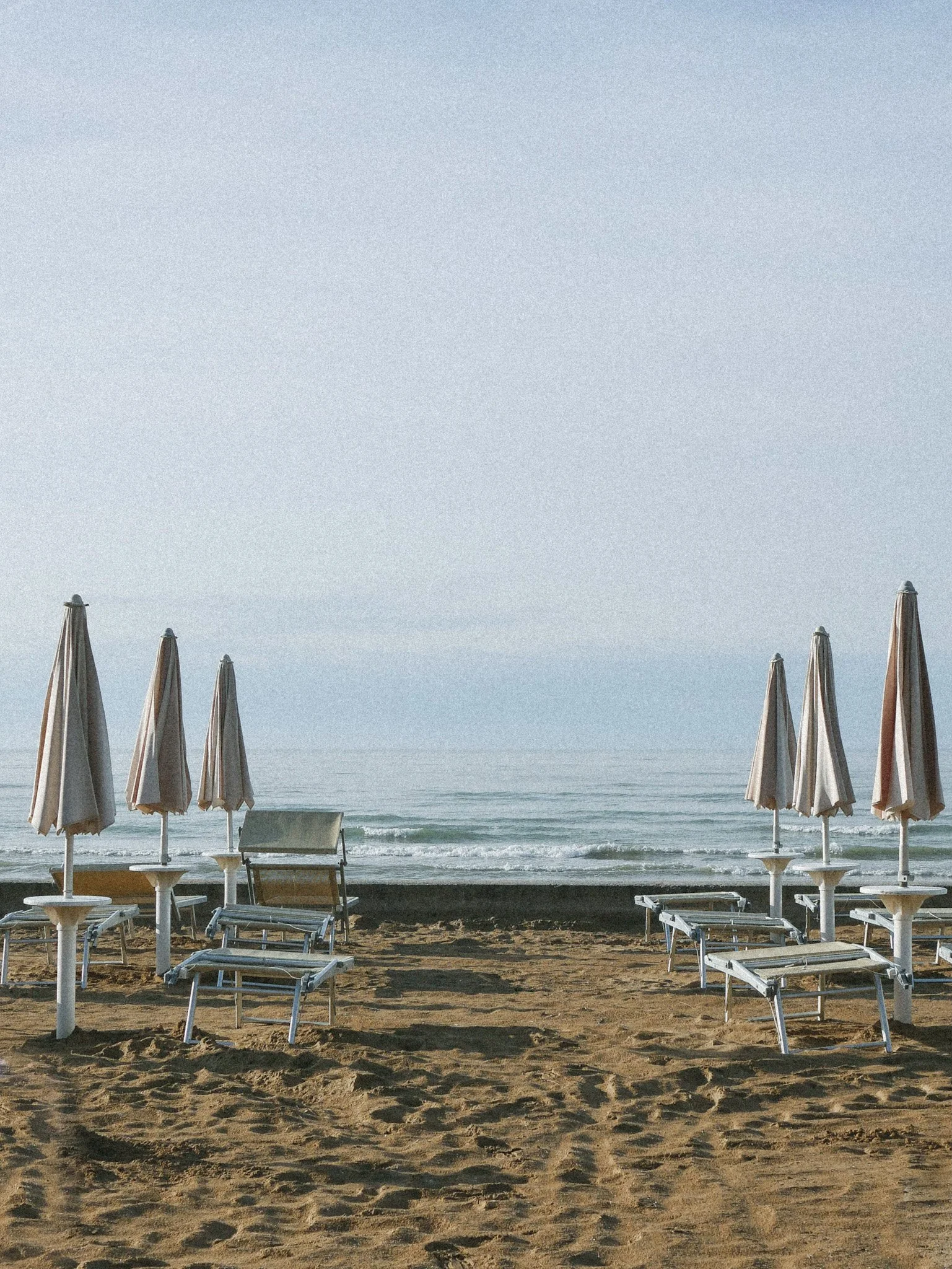 Empty beach with lined-up closed umbrellas and lounge chairs facing the ocean, under a clear sky.