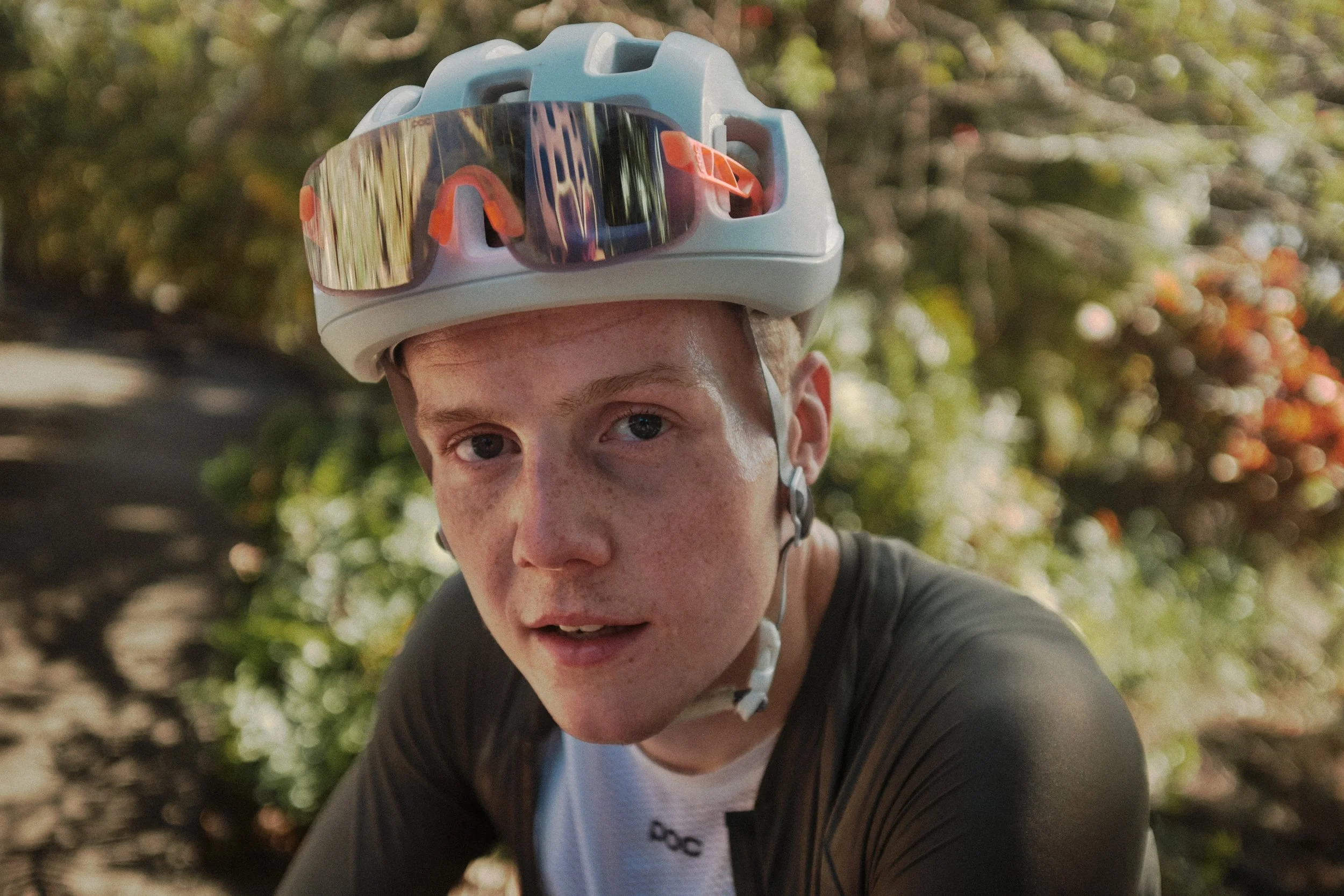 A person with freckles wearing sunglasses and a helmet, outdoors among trees.