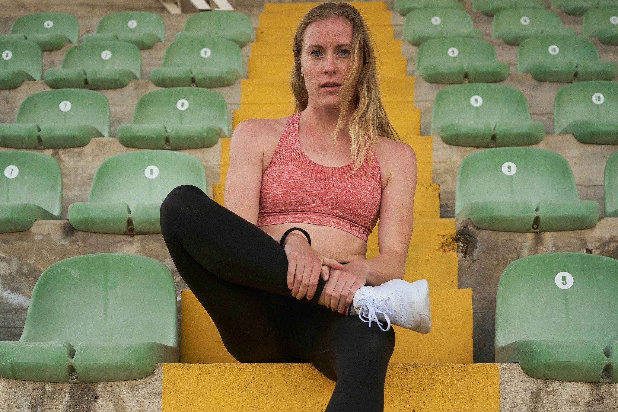 A woman sitting on concrete stadium stairs, with green stadium seats behind her, wearing a red sports bra, black leggings, and white sneakers, with a serious expression.