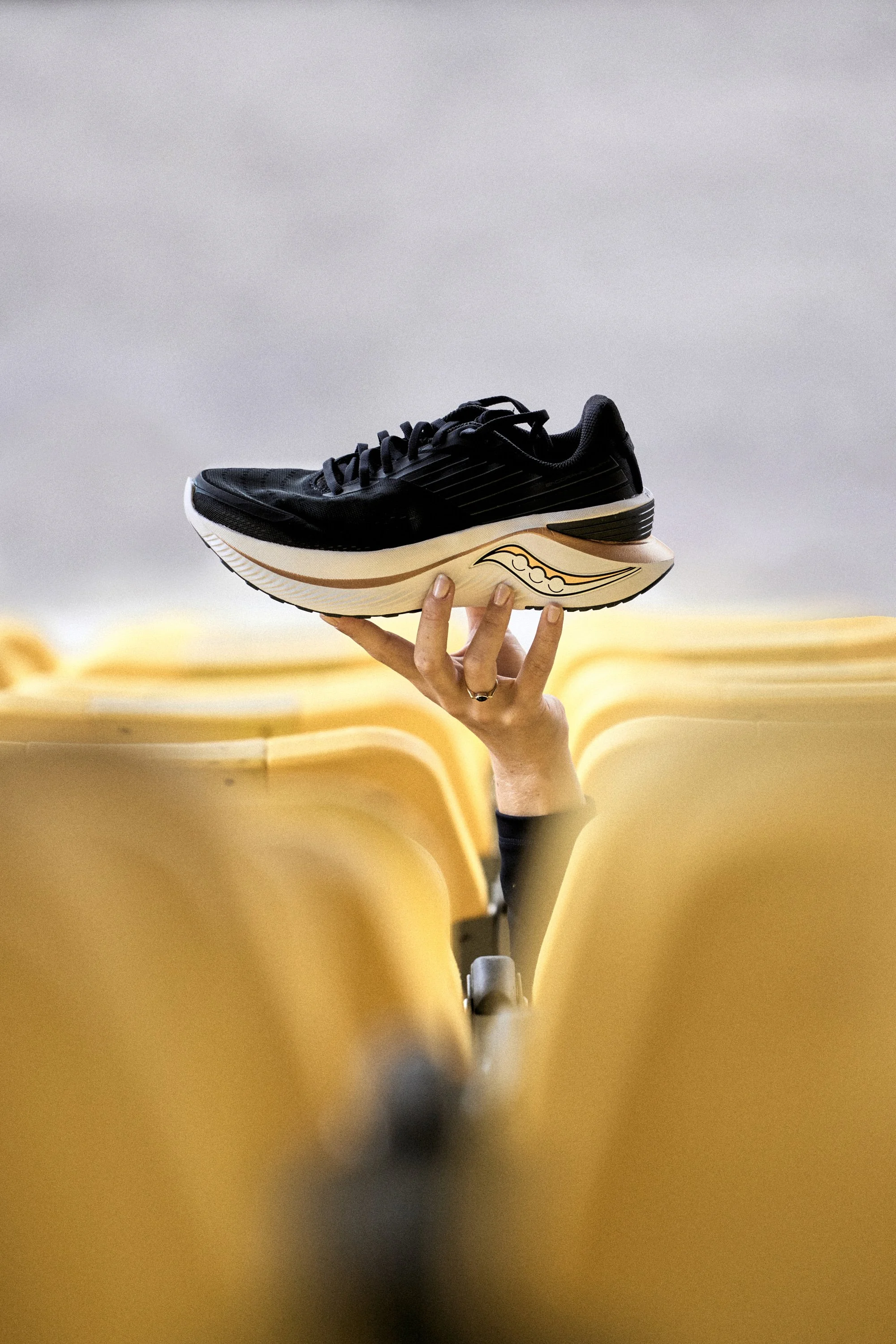 A black athletic shoe with a cream-colored sole being held up by a person's hand in an empty seating area with yellow chairs.