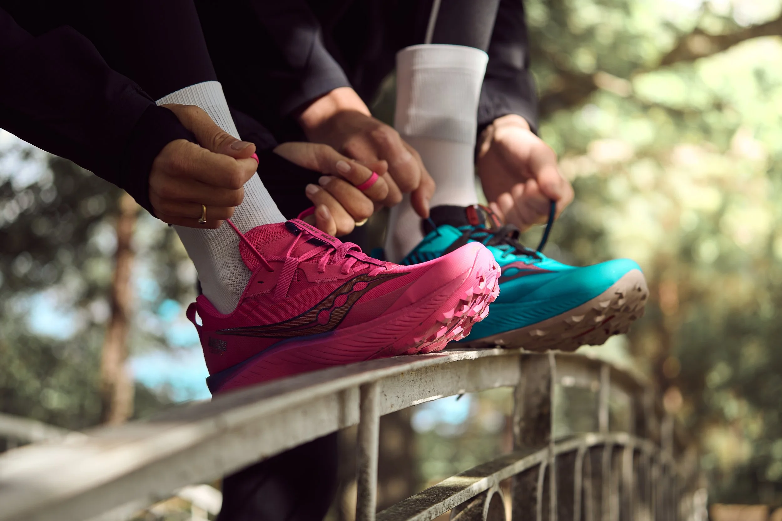 Close-up of two people tying their running shoes, with one person wearing pink shoes and the other wearing teal shoes, on a wooden outdoor structure surrounded by trees.