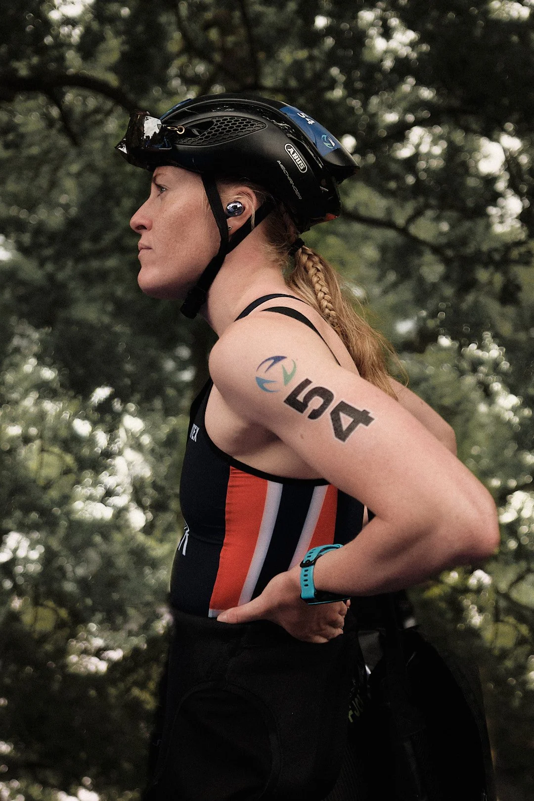 A female athlete wearing a black helmet, sports tank top, and a blue wristband, standing outdoors with a wooded background.
