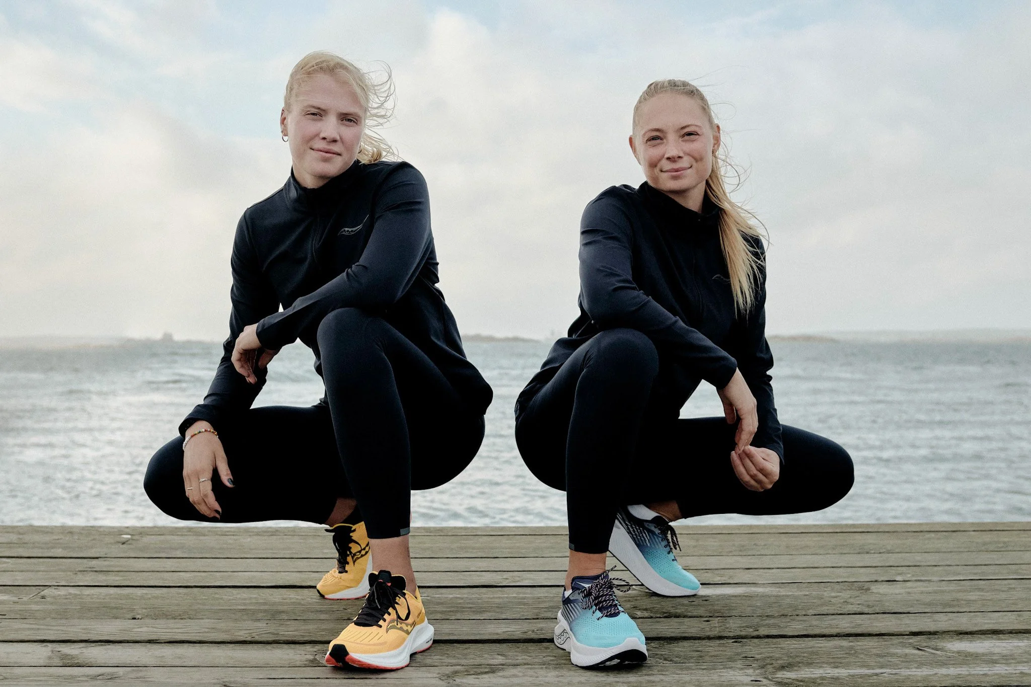 Two young women in athletic clothing squatting on a wooden walkway near the water, with cloudy skies above.