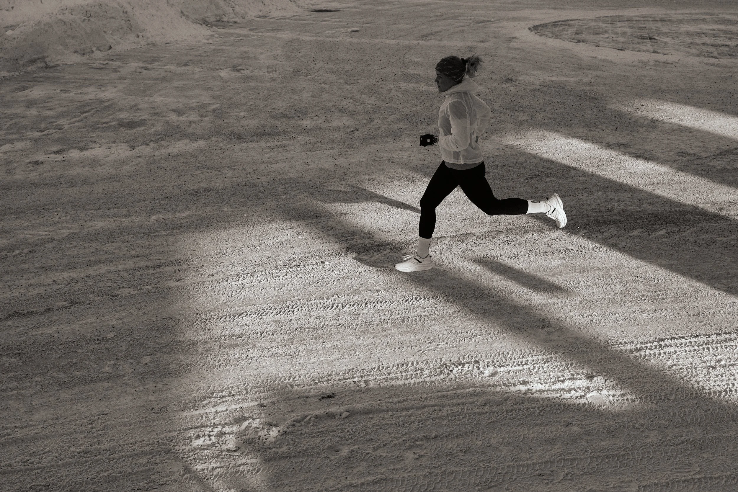 A woman running on a dusty path with tire tracks, wearing athletic gear and headphones.