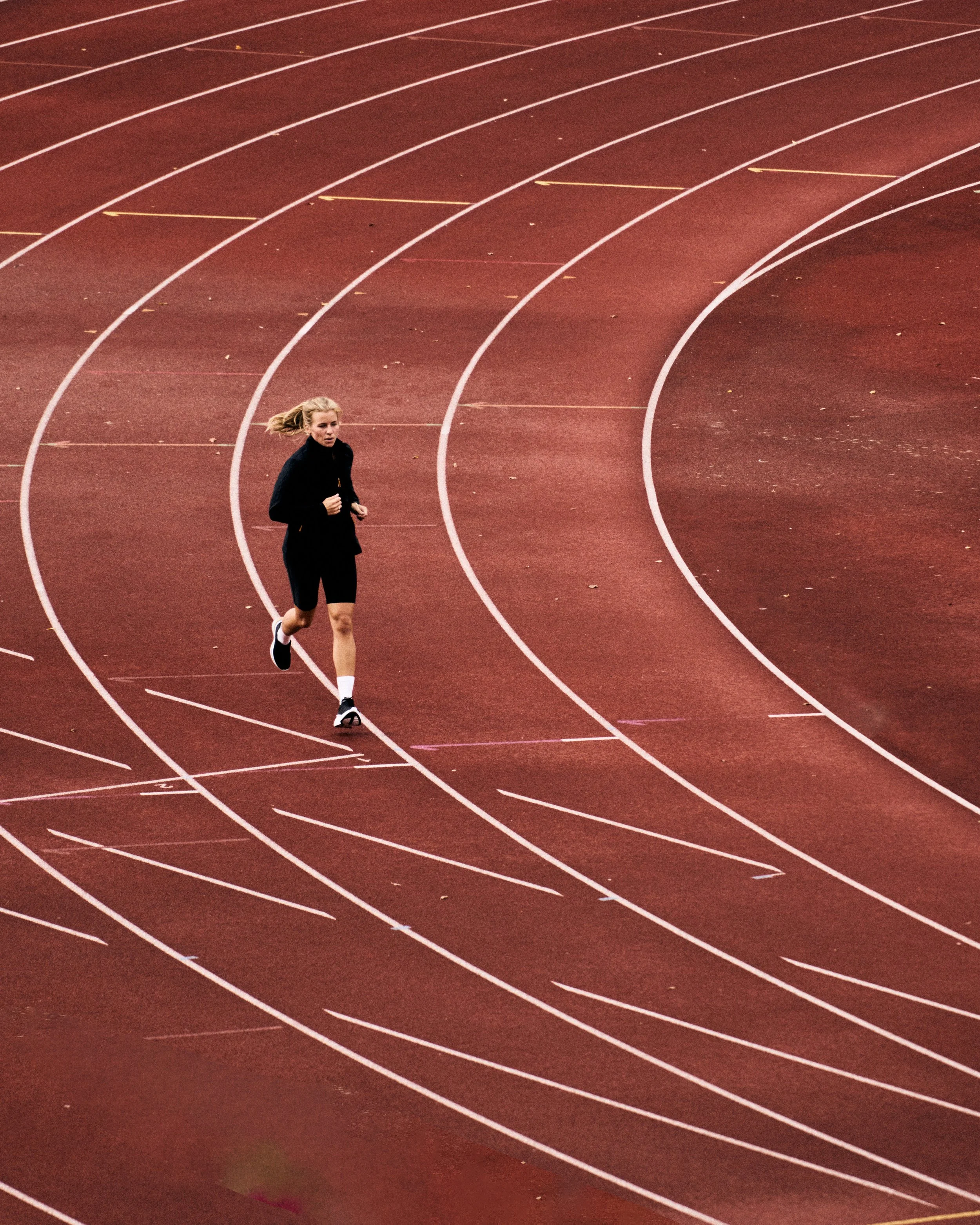 A woman jogging on a red running track with white lane lines, surrounded by scattered leaves.