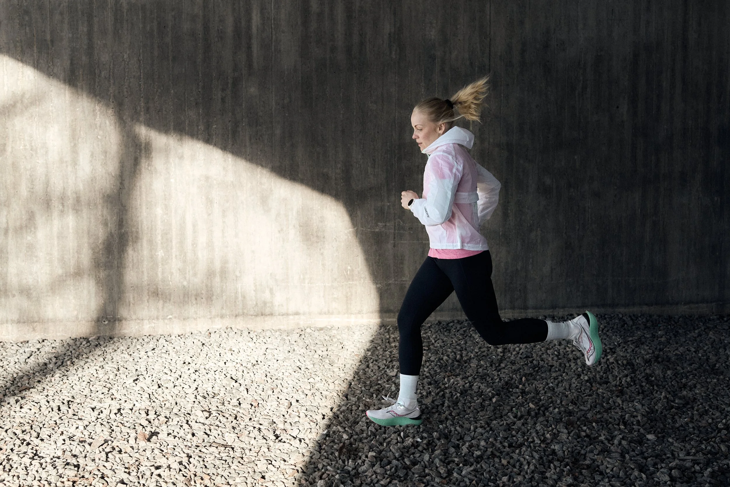 A woman in a pink and white jacket, black leggings, and white running shoes with green soles is jogging outdoors against a dark concrete wall with sunlight casting a shadow on the gravel ground.