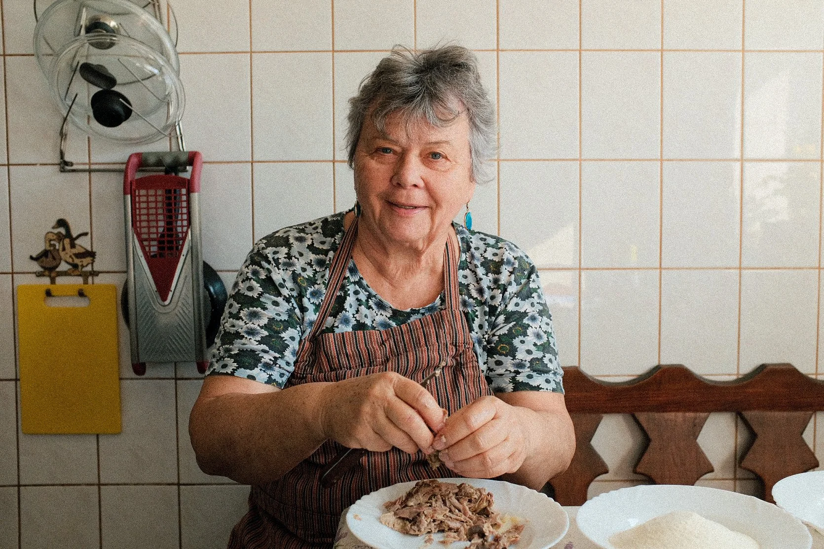 An elderly woman with short gray hair, wearing a floral shirt and striped apron, sits at a table in a kitchen, preparing food with a plate of shredded meat in front of her. The kitchen has white tiled walls, a yellow cutting board, and hanging kitche