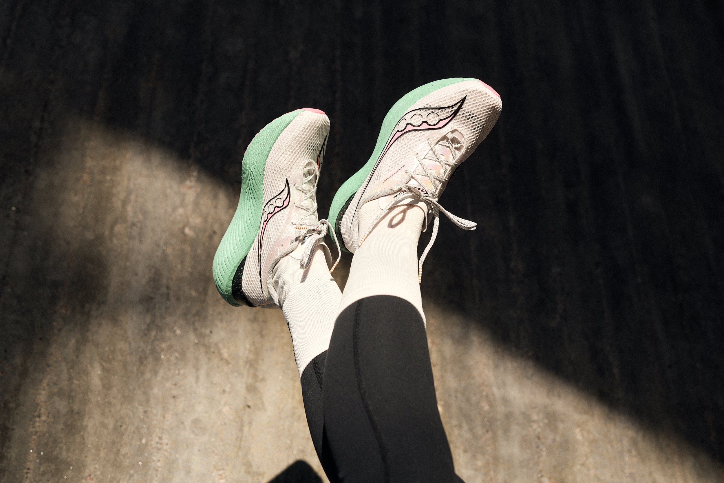 A person wearing white running shoes with green soles and black and white striped leggings, standing on a dark concrete floor.
