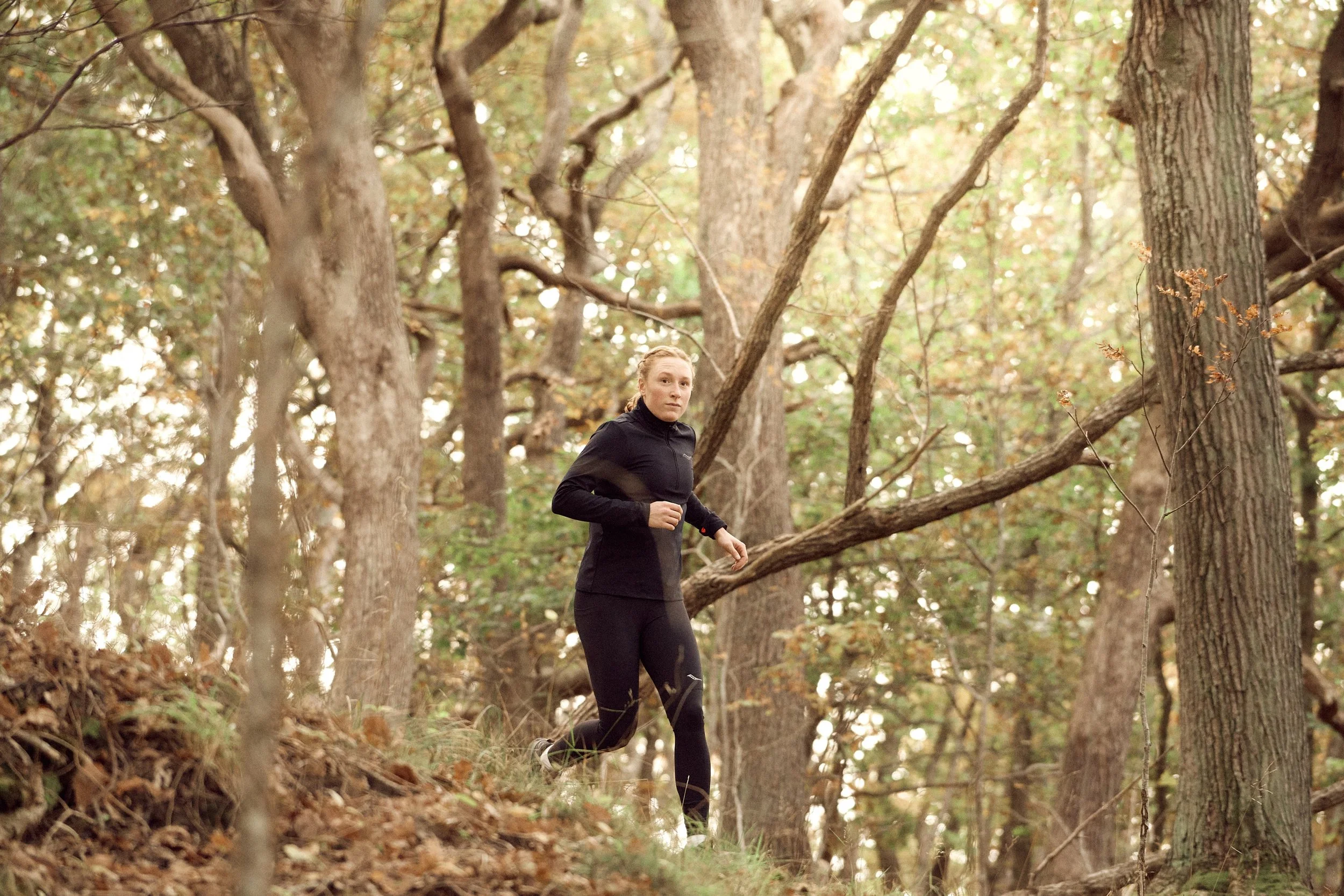 A woman jogging through a forested trail during daytime, surrounded by tall trees with green and brown leaves.