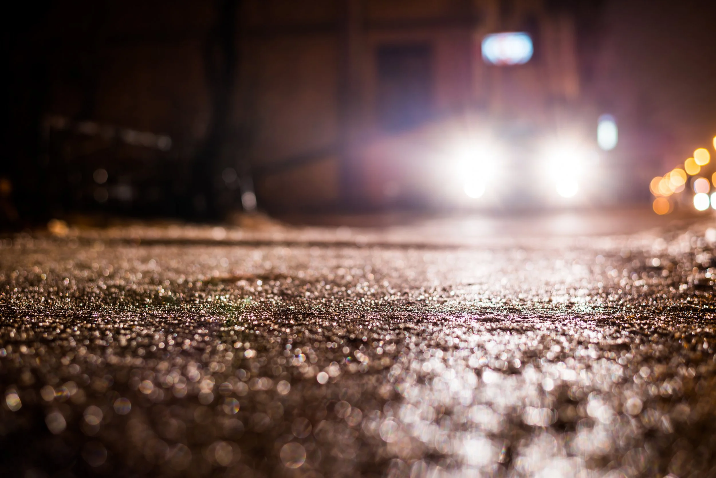 Close-up of a wet city street at night with bright headlights in the distance and blurred lights on the right side.