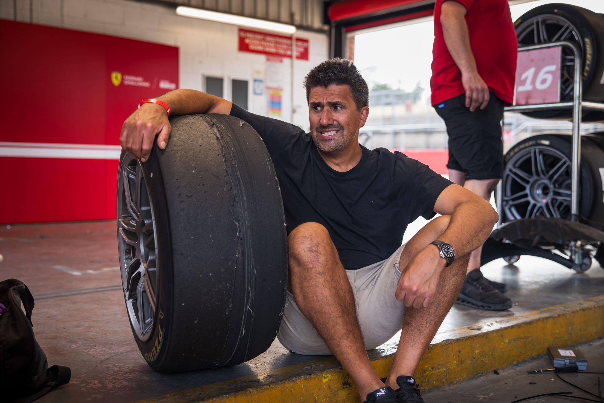 John Marcar sitting on the ground next to a worn racing tire in a garage, appearing distressed or exhausted.