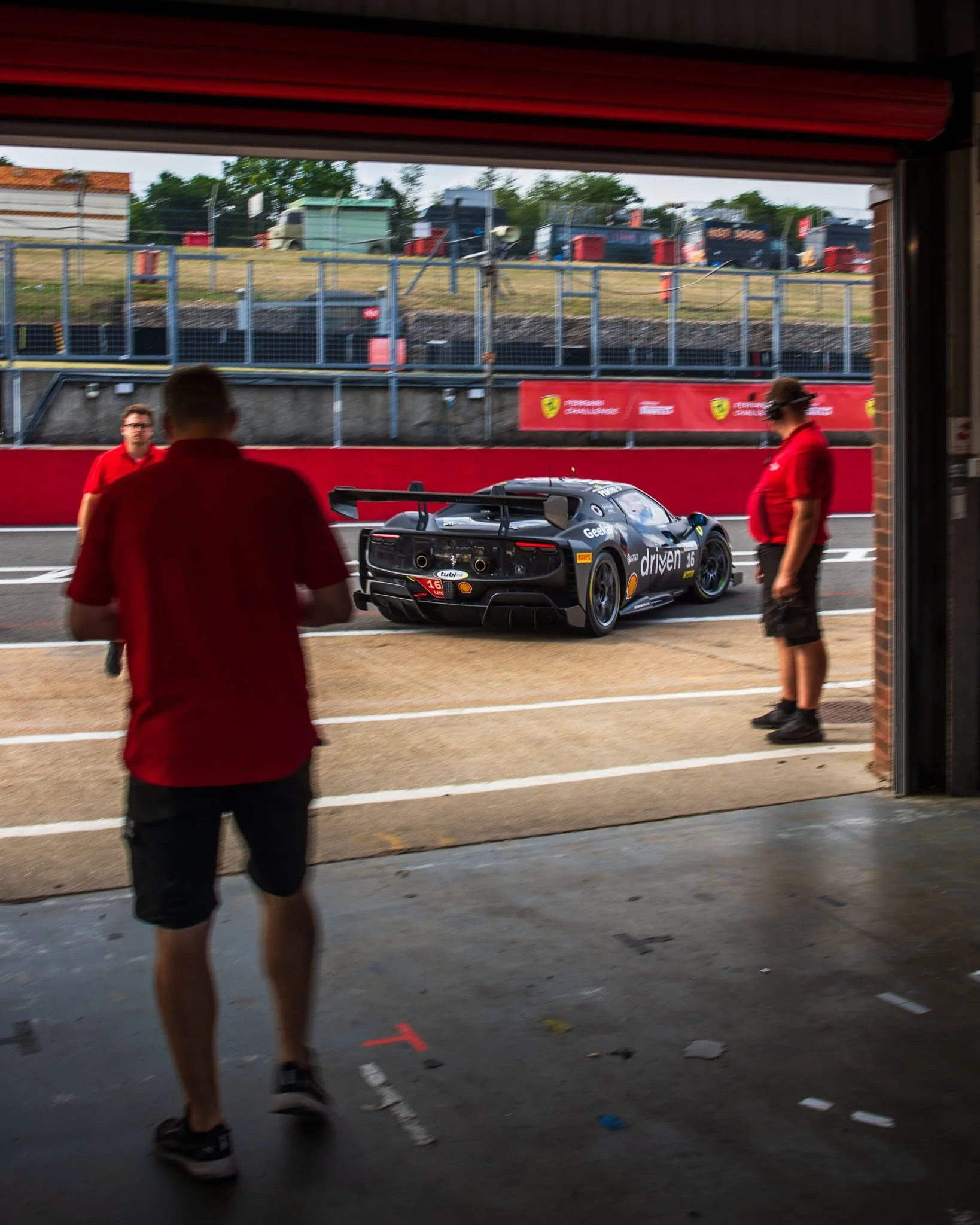 A race car in the pit lane at the race track, with team members standing nearby, view seen from inside a garage.