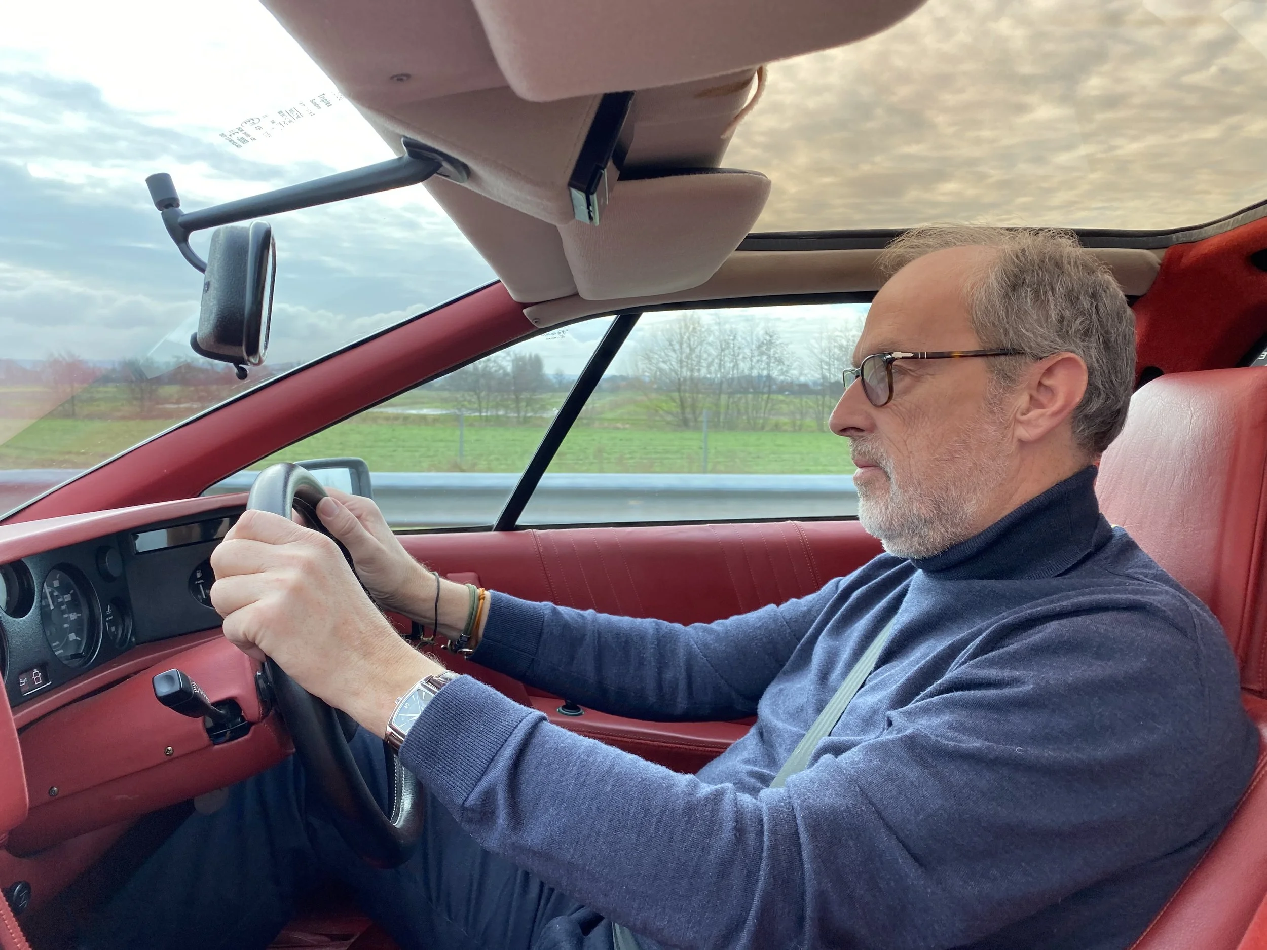 A middle-aged man with glasses and gray hair driving a car with red interior on a cloudy day.