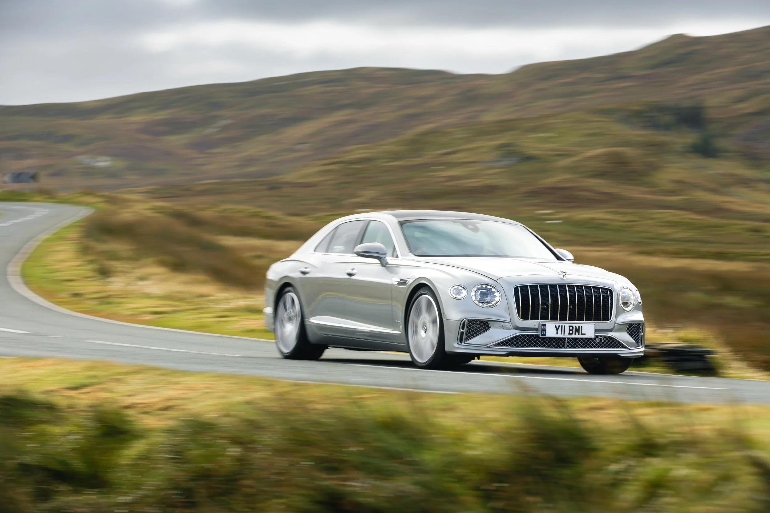 A silver luxury sedan driving on a winding road through a hilly landscape under a cloudy sky.