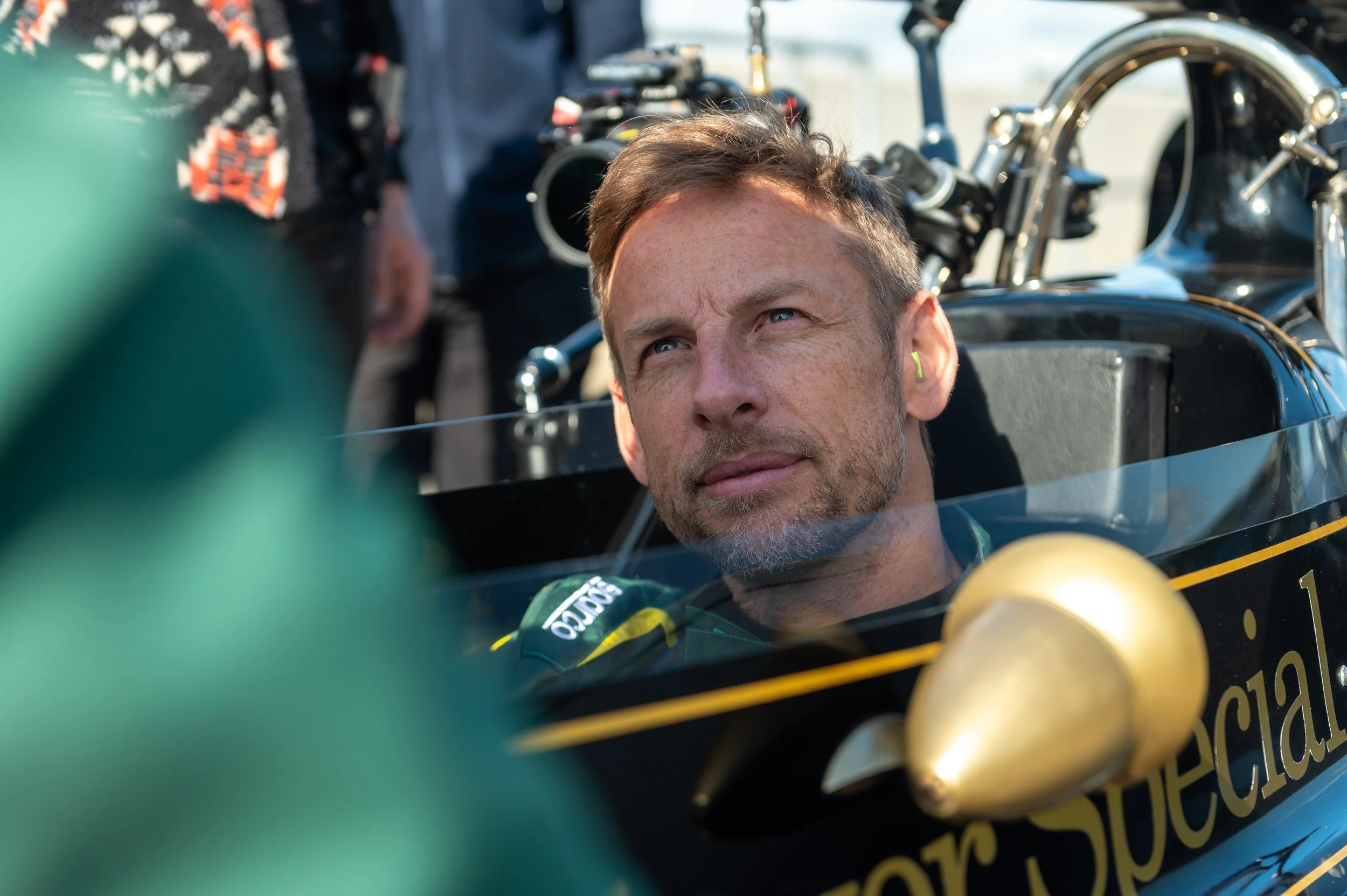 A man with short blonde hair and a beard sitting in a vintage black race car with gold accents, looking at someone outside the car. In the background, there are other people and racing equipment.