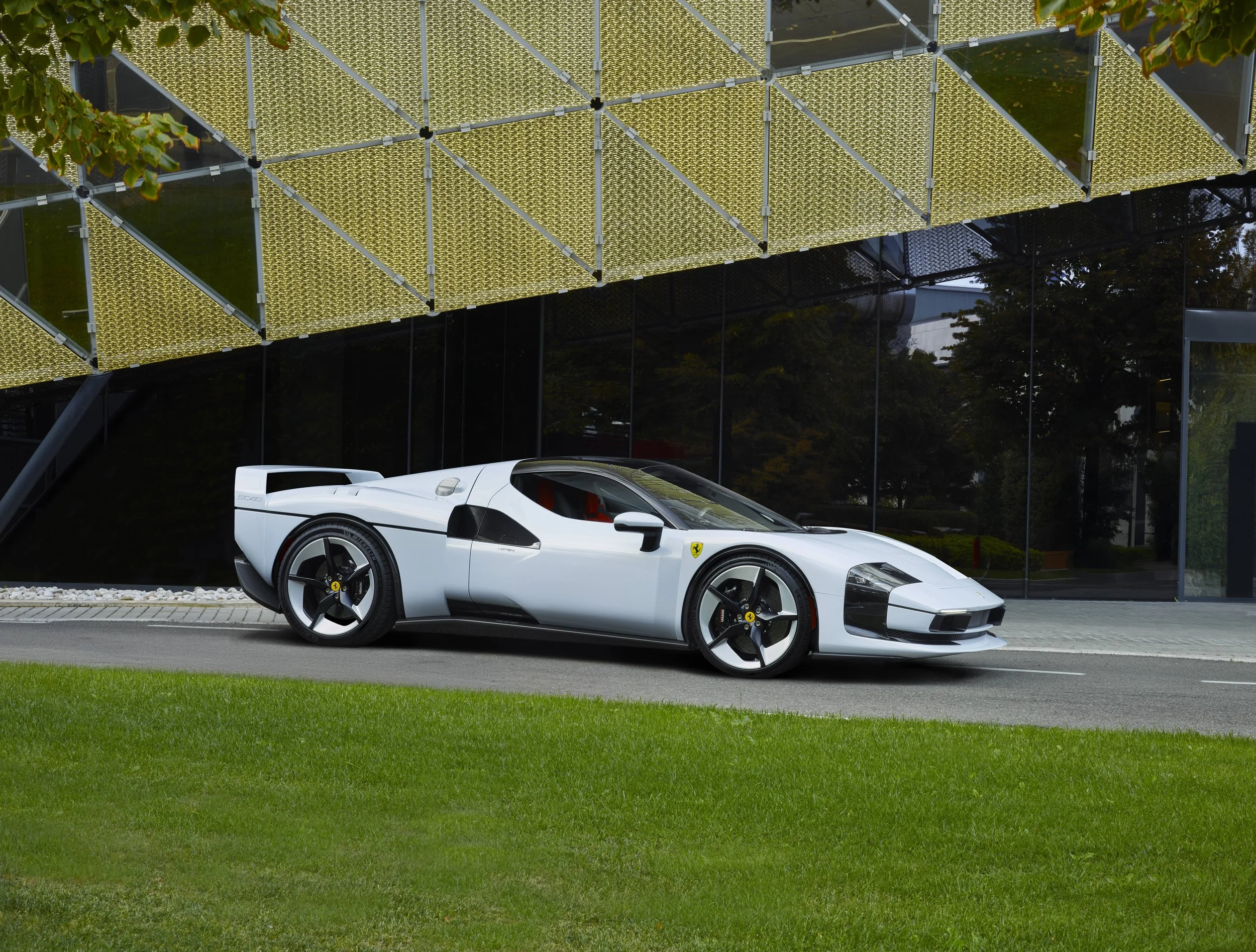 A white Ferrari sports car parked in front of a modern glass building with a gold-colored facade.