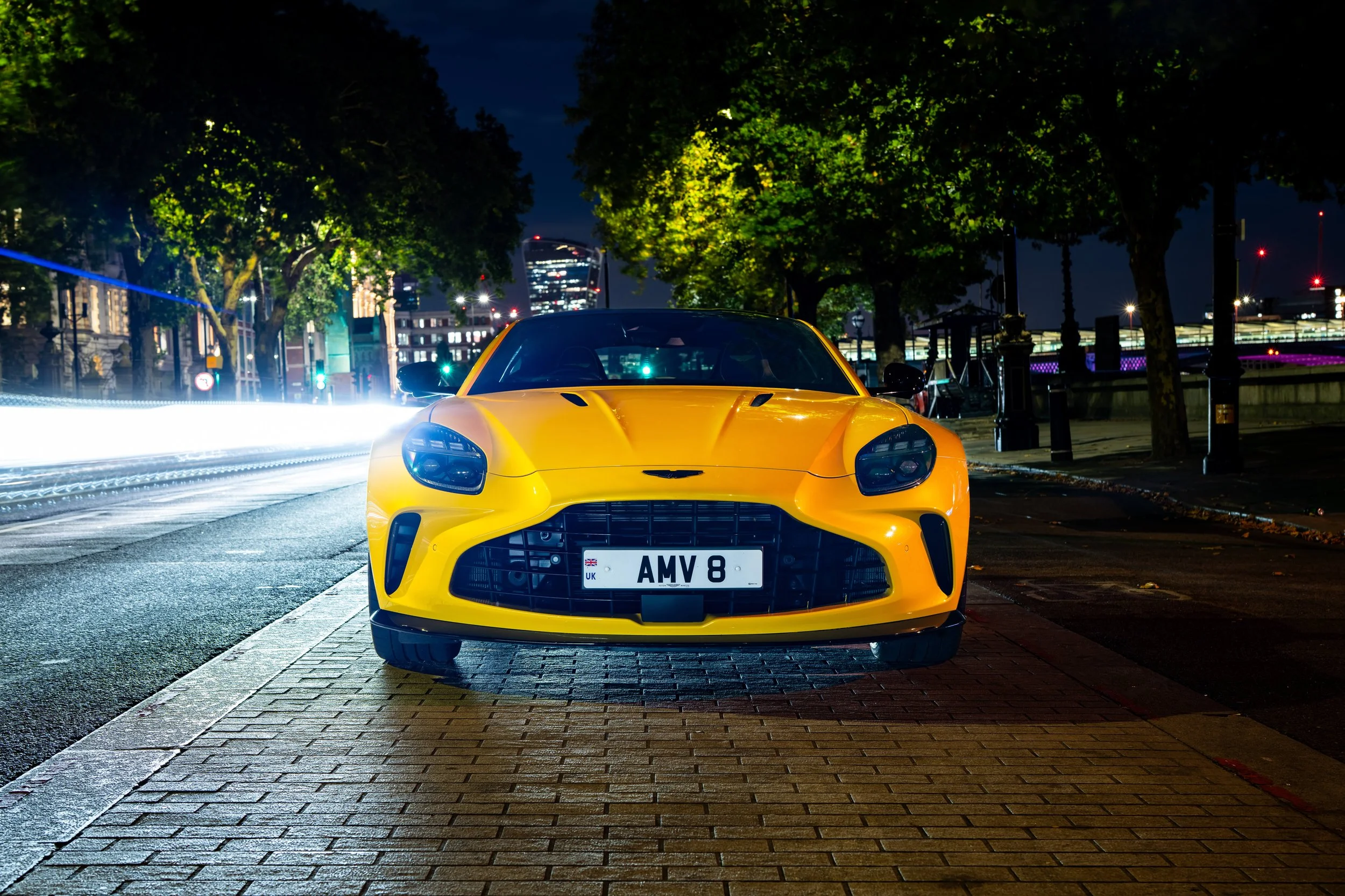 A yellow sports car parked on a city street at night, with city lights and trees in the background.