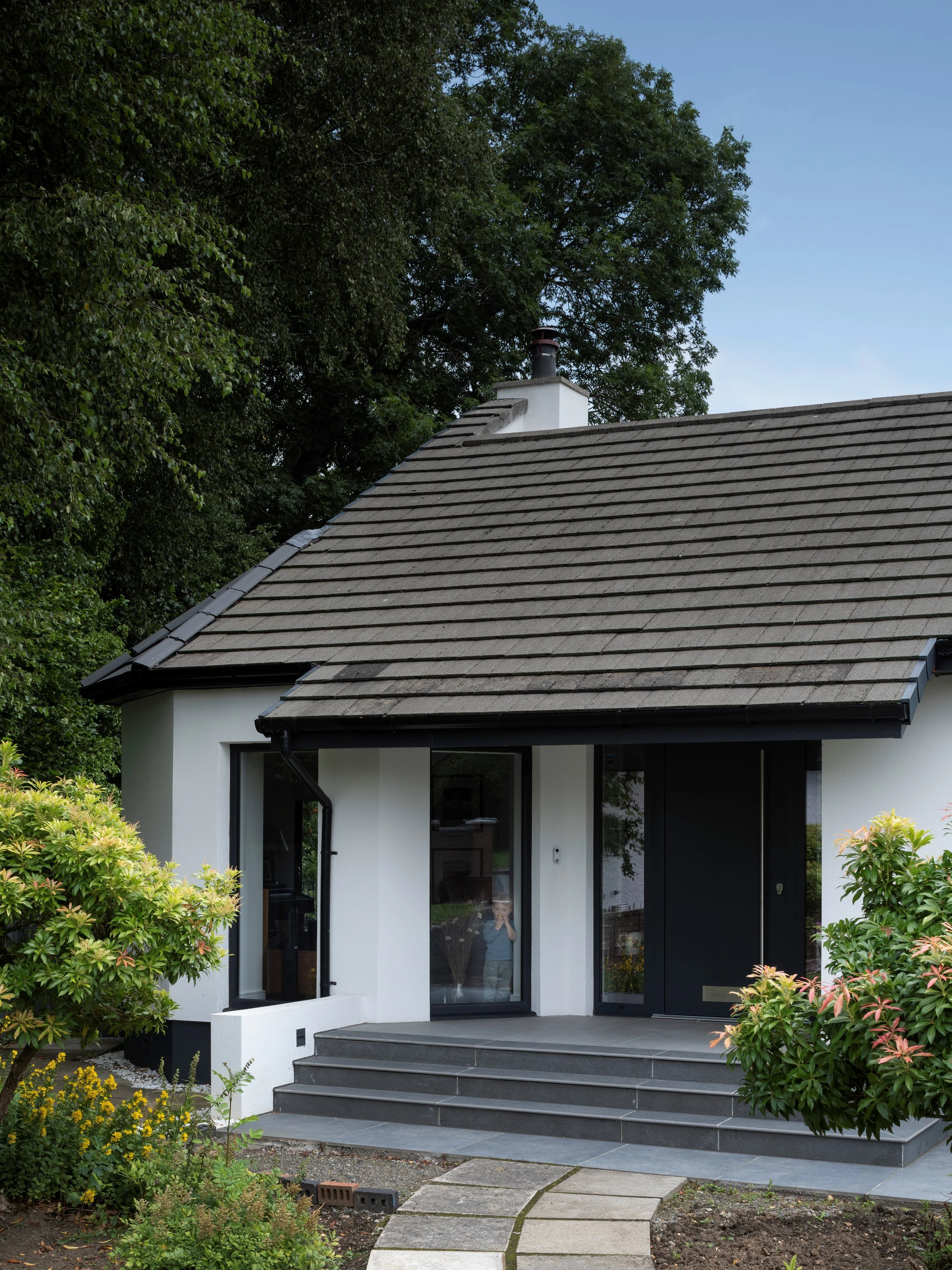 A modern house with black-framed glass doors, a gray tiled sloped roof, surrounded by green bushes and trees, with a stone pathway leading to the entrance and a person taking a picture in the reflection of the glass door.