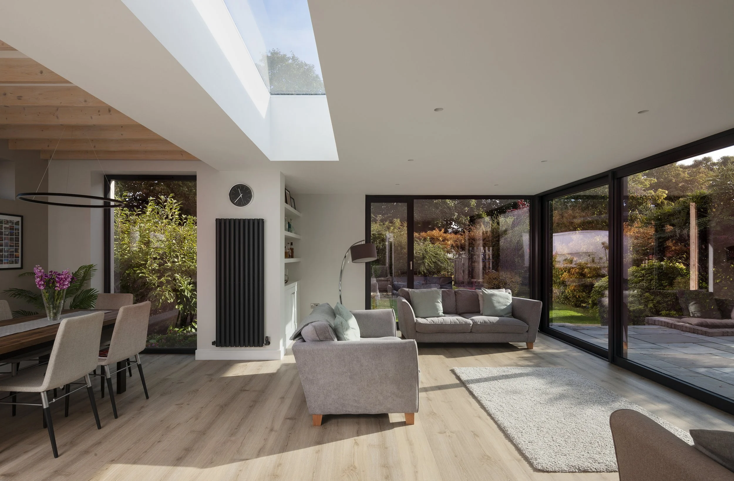 A modern living room with large glass windows and sliding doors showing an outdoor garden with trees and shrubs. The room has a skylight, a white ceiling, light-colored hardwood floors, a gray sofa, a black floor lamp, and a white wall with a black c