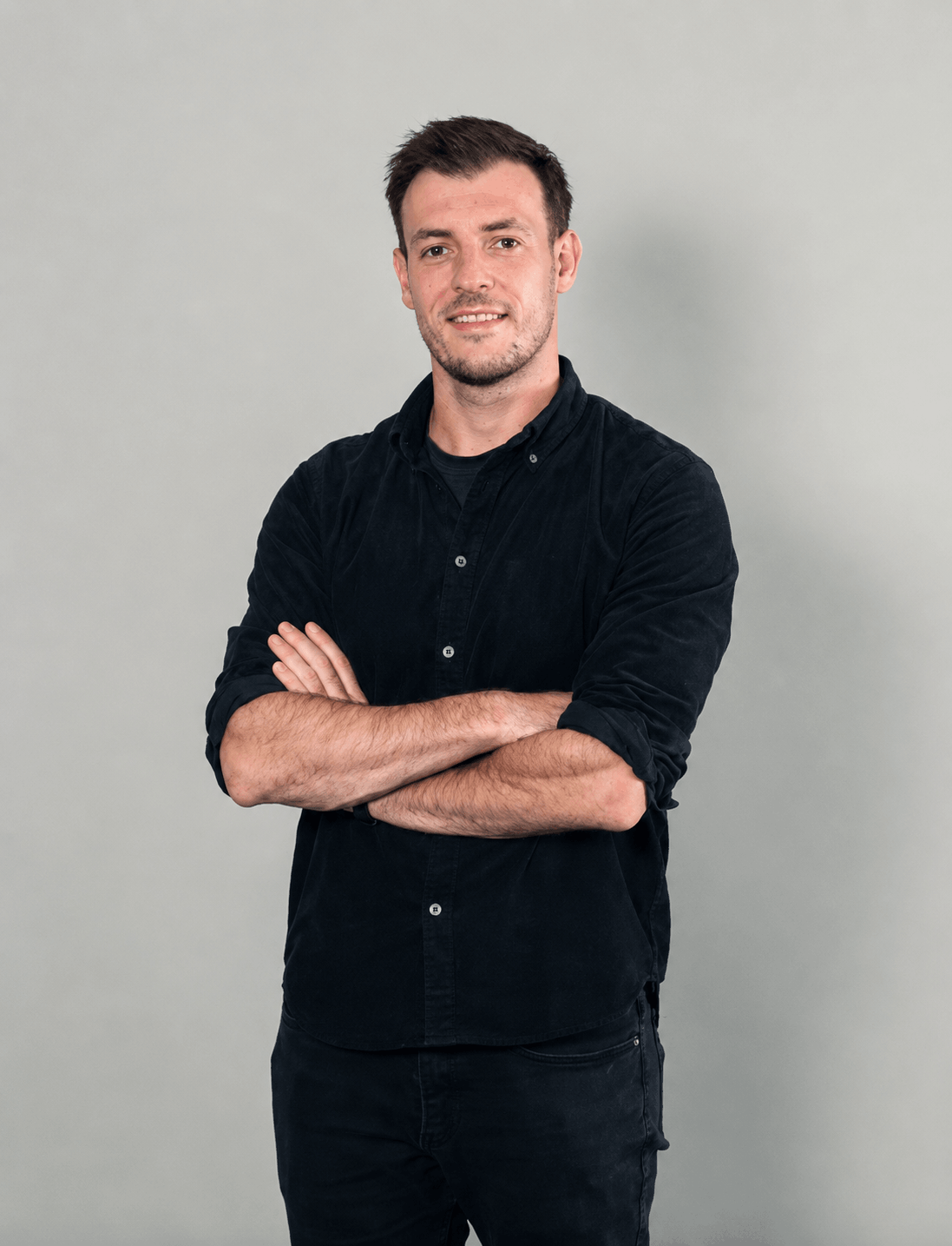 A young man with short dark hair and a beard, wearing a black button-up shirt with sleeves rolled up, standing with arms crossed against a plain light gray wall.