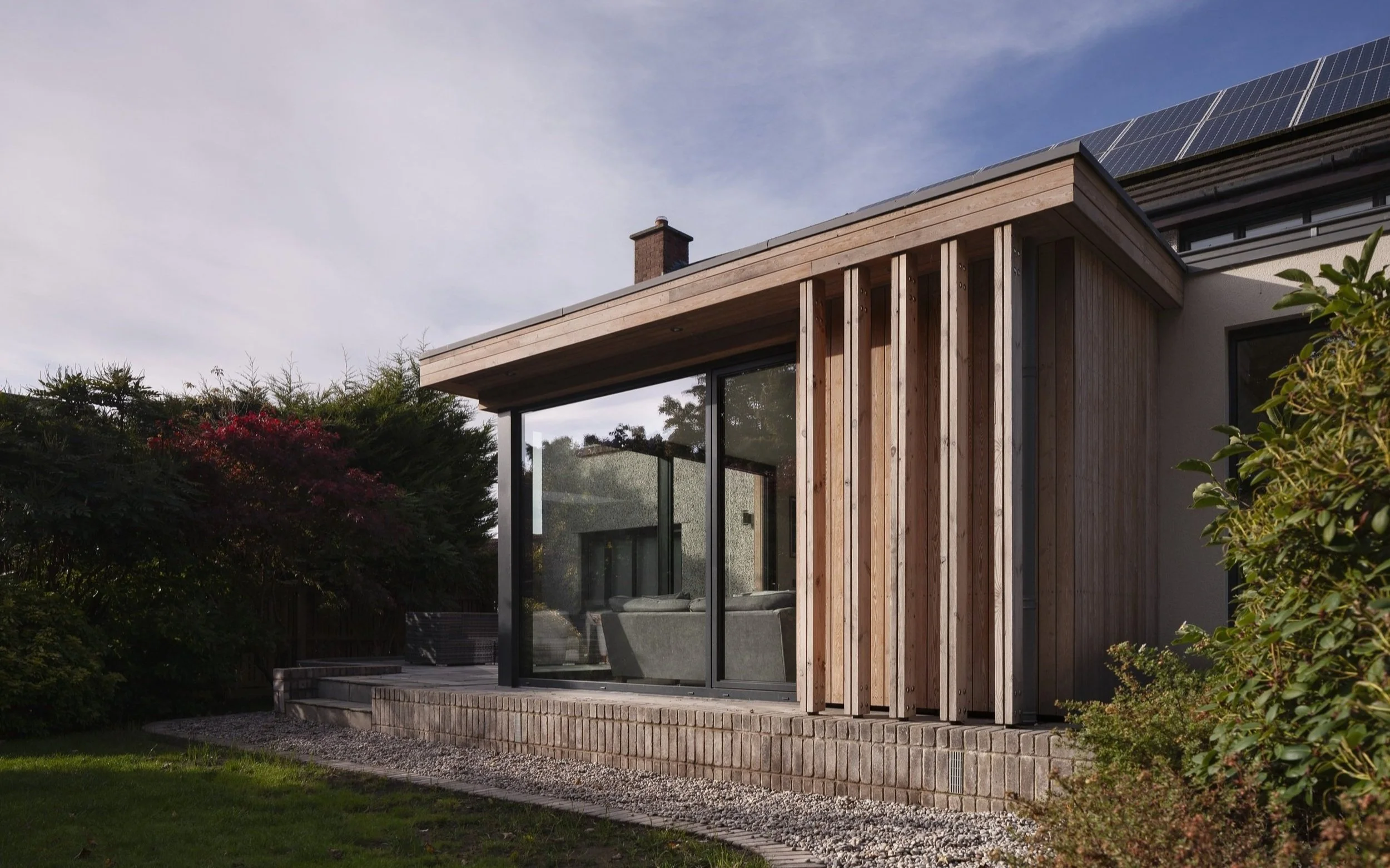 A modern house with wooden paneling, glass sliding doors, and solar panels on the roof, surrounded by greenery and a landscaped yard.