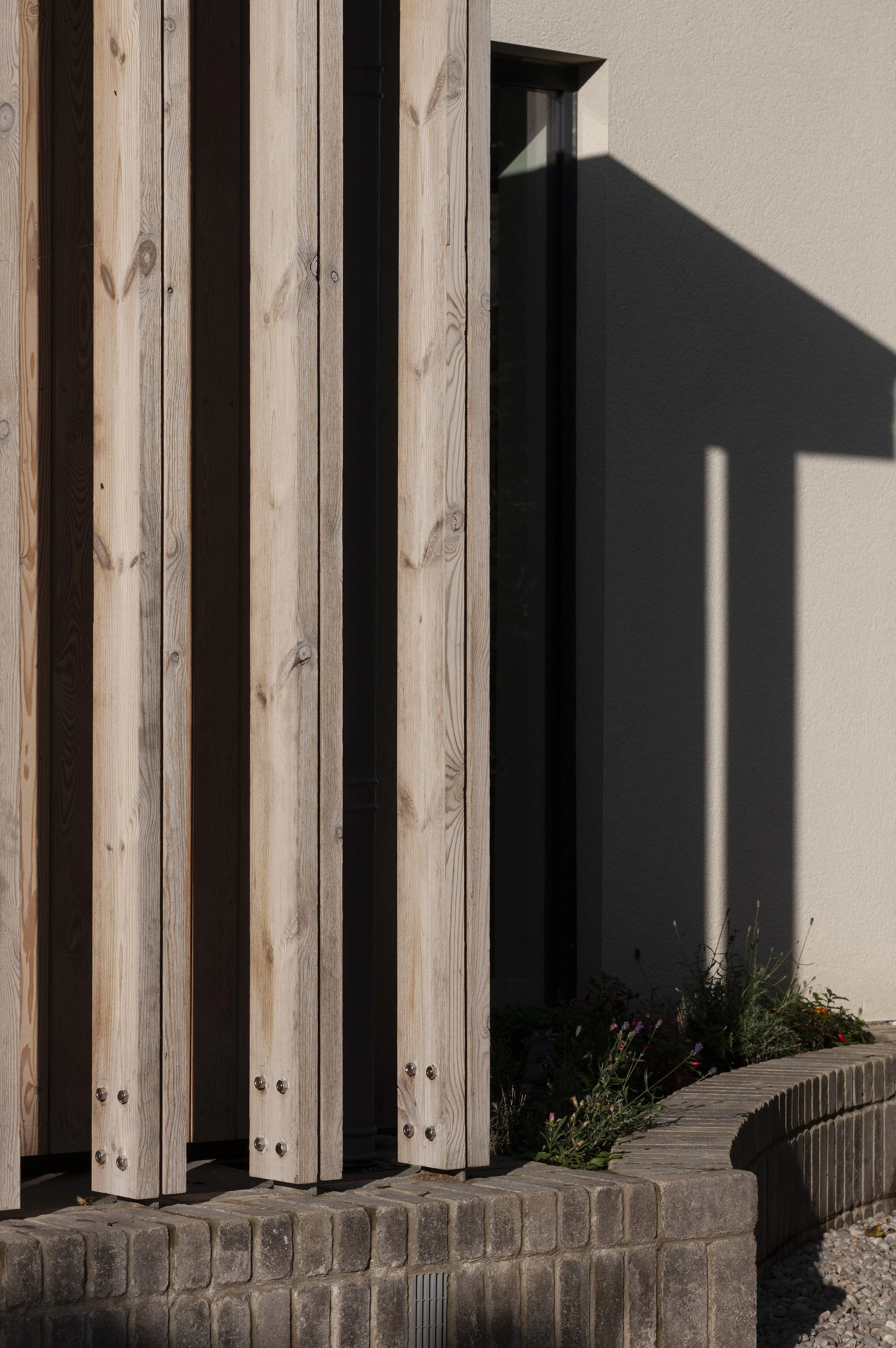 Close-up photo of a modern building exterior with vertical wooden slats, a small narrow window, a gray concrete wall, plants, and shadows cast by a structure.