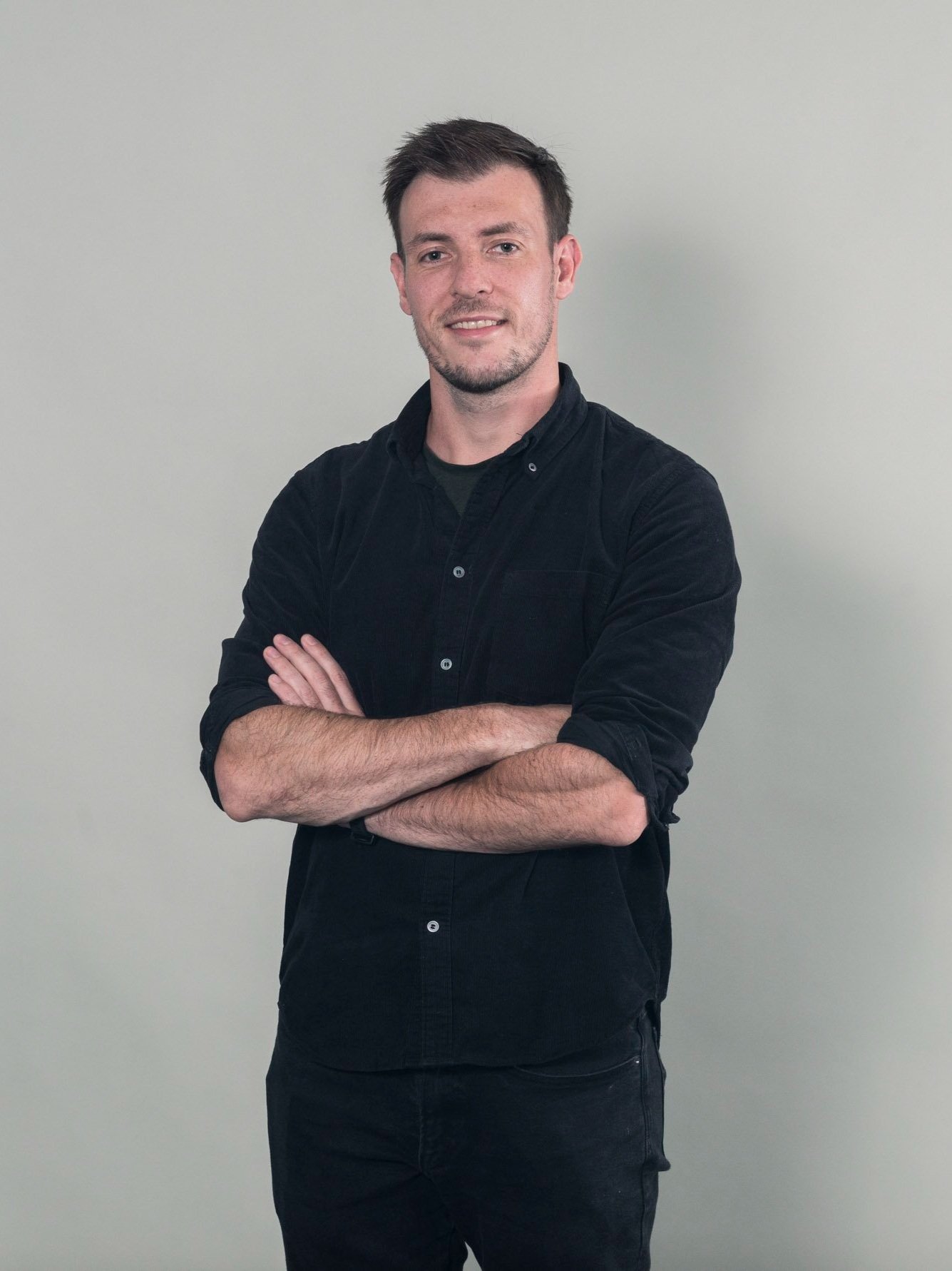 A young man with dark hair and a beard, smiling with arms crossed, wearing a black button-up shirt and black pants, standing against a light gray background.