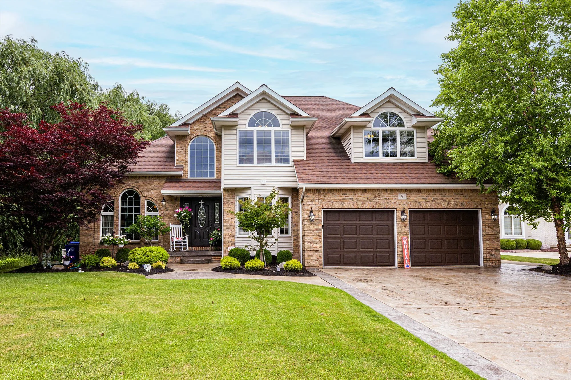 Front view of a two-story brick and siding house with a two-car garage, lush front lawn, and decorative landscaping, under a partly cloudy sky.
