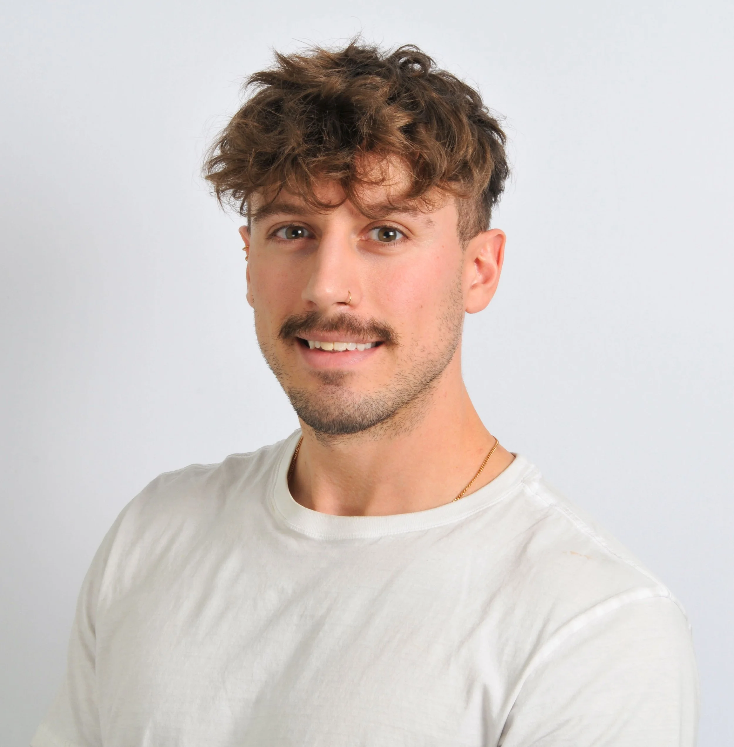 Portrait of a young man with curly brown hair, light facial hair, wearing a white t-shirt, smiling, against a plain light background.
