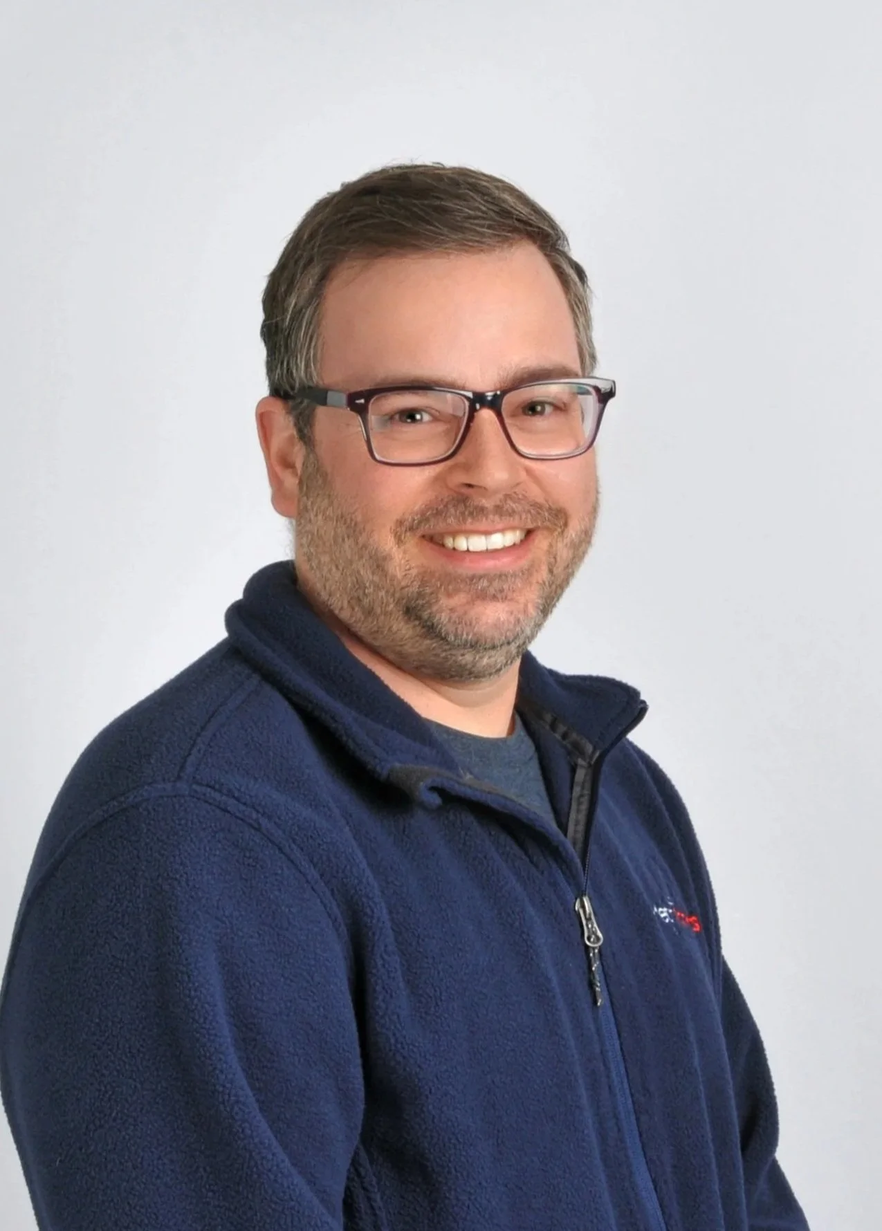 A smiling man with glasses and brown hair, wearing a navy blue fleece jacket, standing against a plain white background.