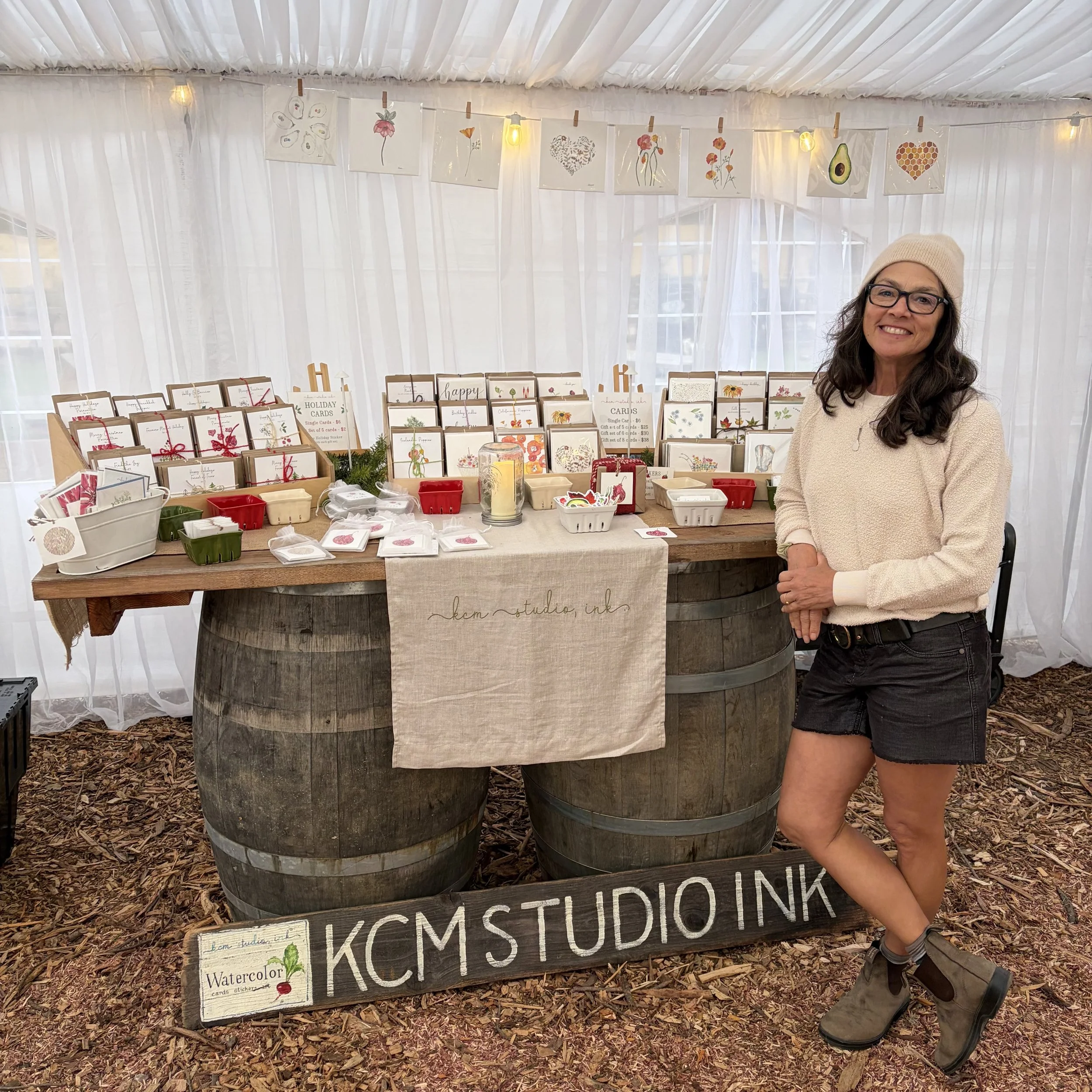 A woman standing next to a display table of greeting cards and watercolor artwork at an indoor craft fair. The table is made from two large barrels and decorated with a beige cloth and a black wood sign reading 'KCMSTUDIO INK'. The background features white curtains with hanging artwork and string lights.