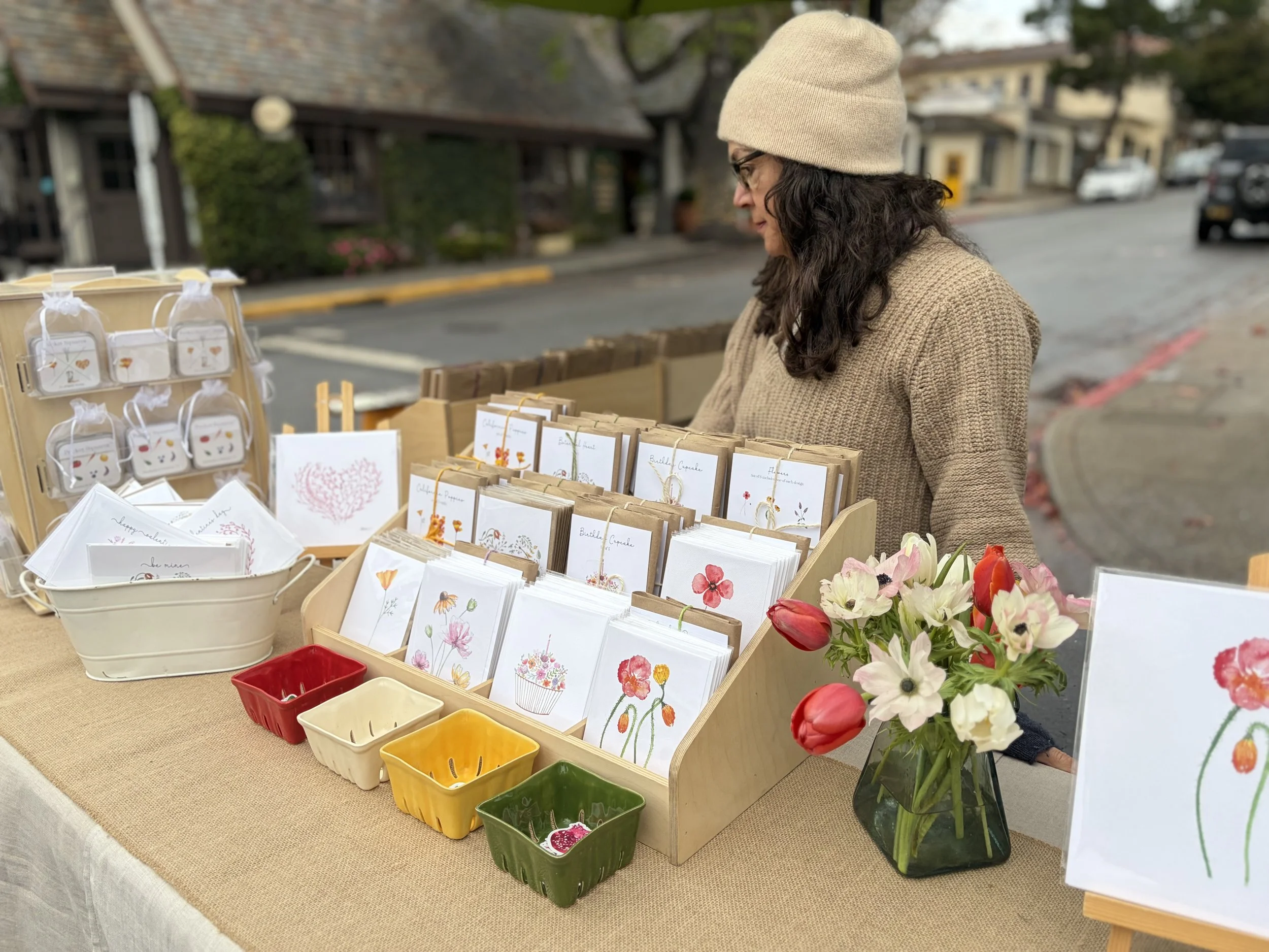 A woman with curly dark hair wearing a beige knit beanie and a matching sweater, standing at an outdoor market stall that displays greeting cards with floral designs, small decorative items, and a bouquet of mixed flowers in a black vase.