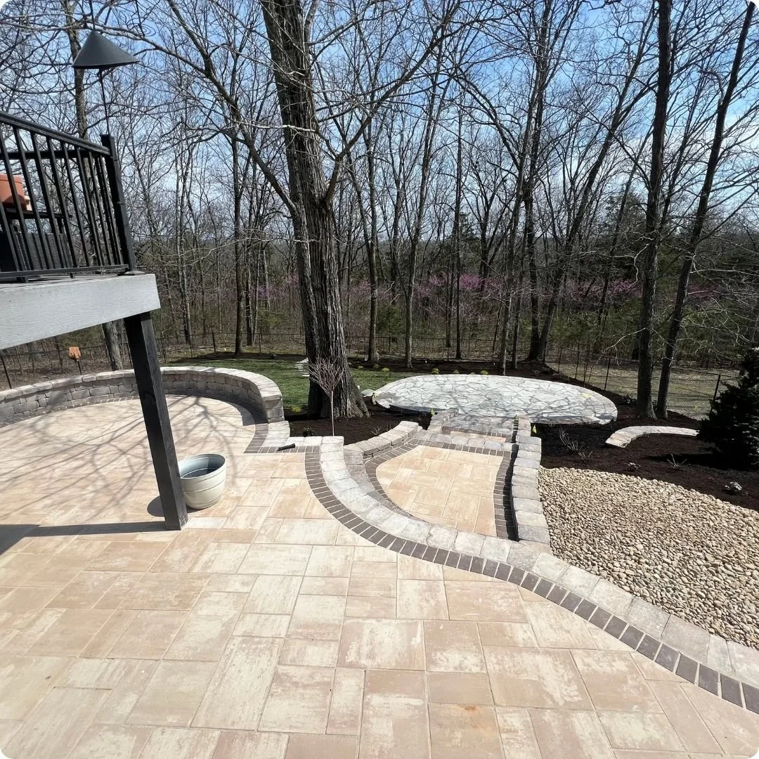 Multi-level paver patio installation featuring Belgard pavers, contrasting dark border inlay, curved natural stone seating wall, and gravel transition beds. A sloped backyard turned into a fully engineered, high-end outdoor living space. Custom hards