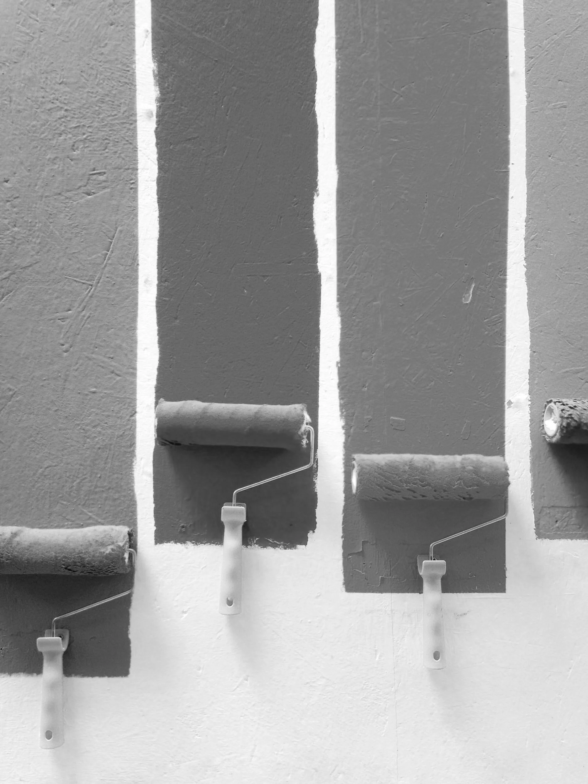Four paint rollers with handles are used to paint a wall, with three rollers showing painted dark grey and one roller on the right with faint paint.