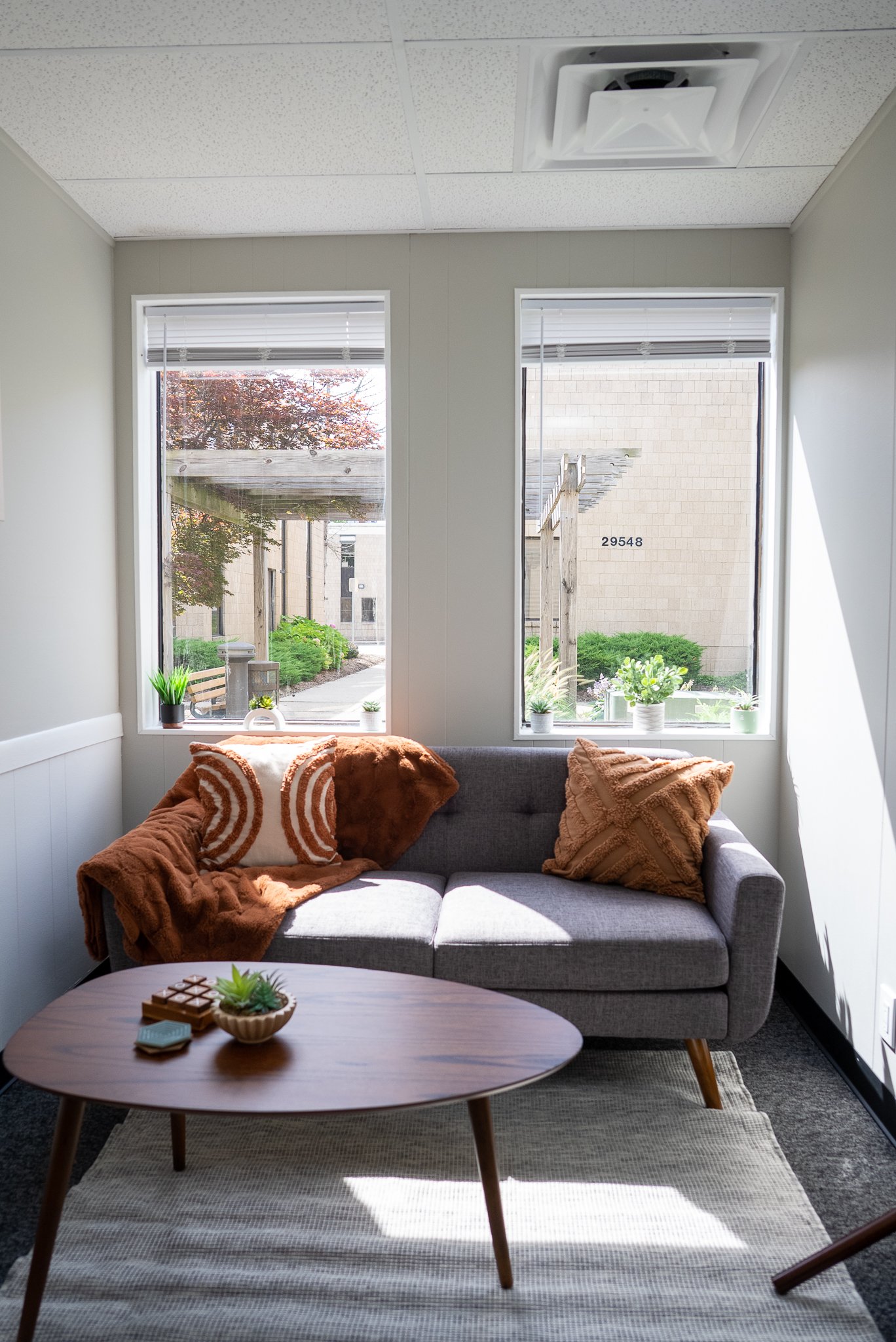 therapy room with gray sofa, orange and beige throw pillows, wooden coffee table with decor, area rug, and large windows showing outdoor garden with trees, plants, and building exterior. EFT Couples Therapy in Royal Oak, Michigan and online statewide