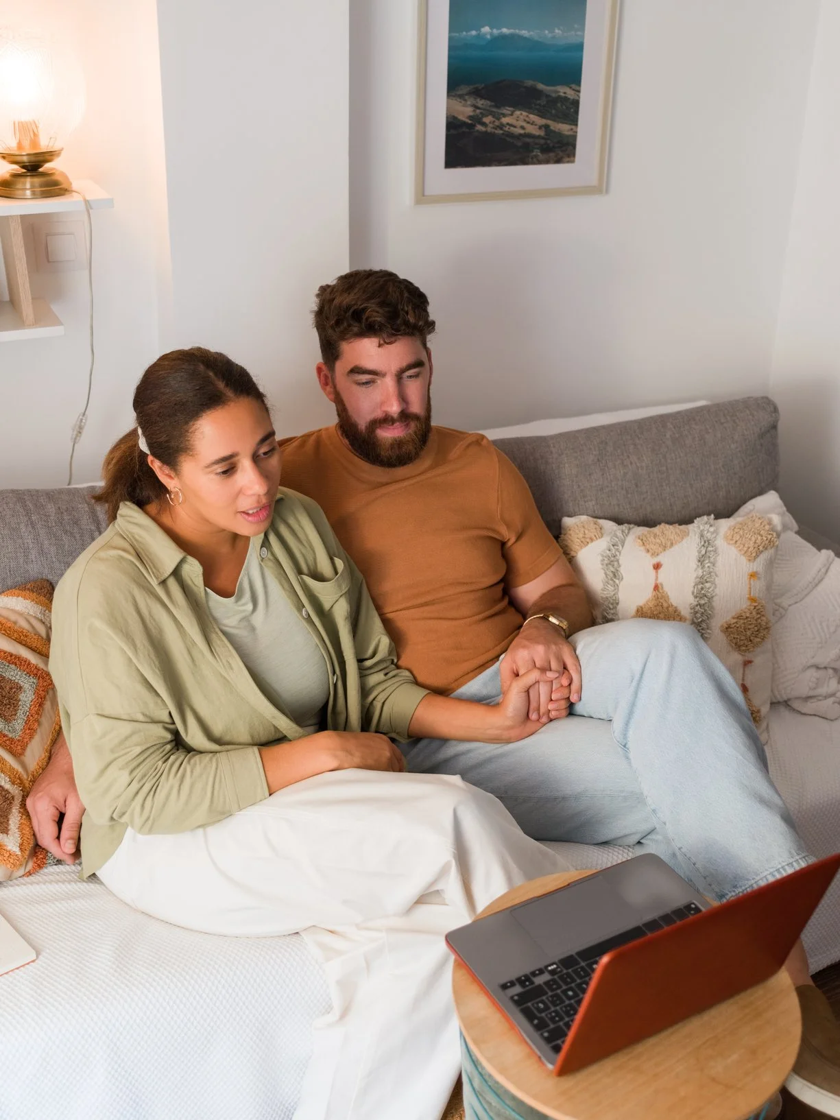 A man and woman sitting on a bed holding hands and watching a laptop screen, with a wooden table holding the laptop in front of them, a gray sofa, and a photograph on the white wall behind them. EFT Couples Therapy in Royal Oak, Michigan and online.