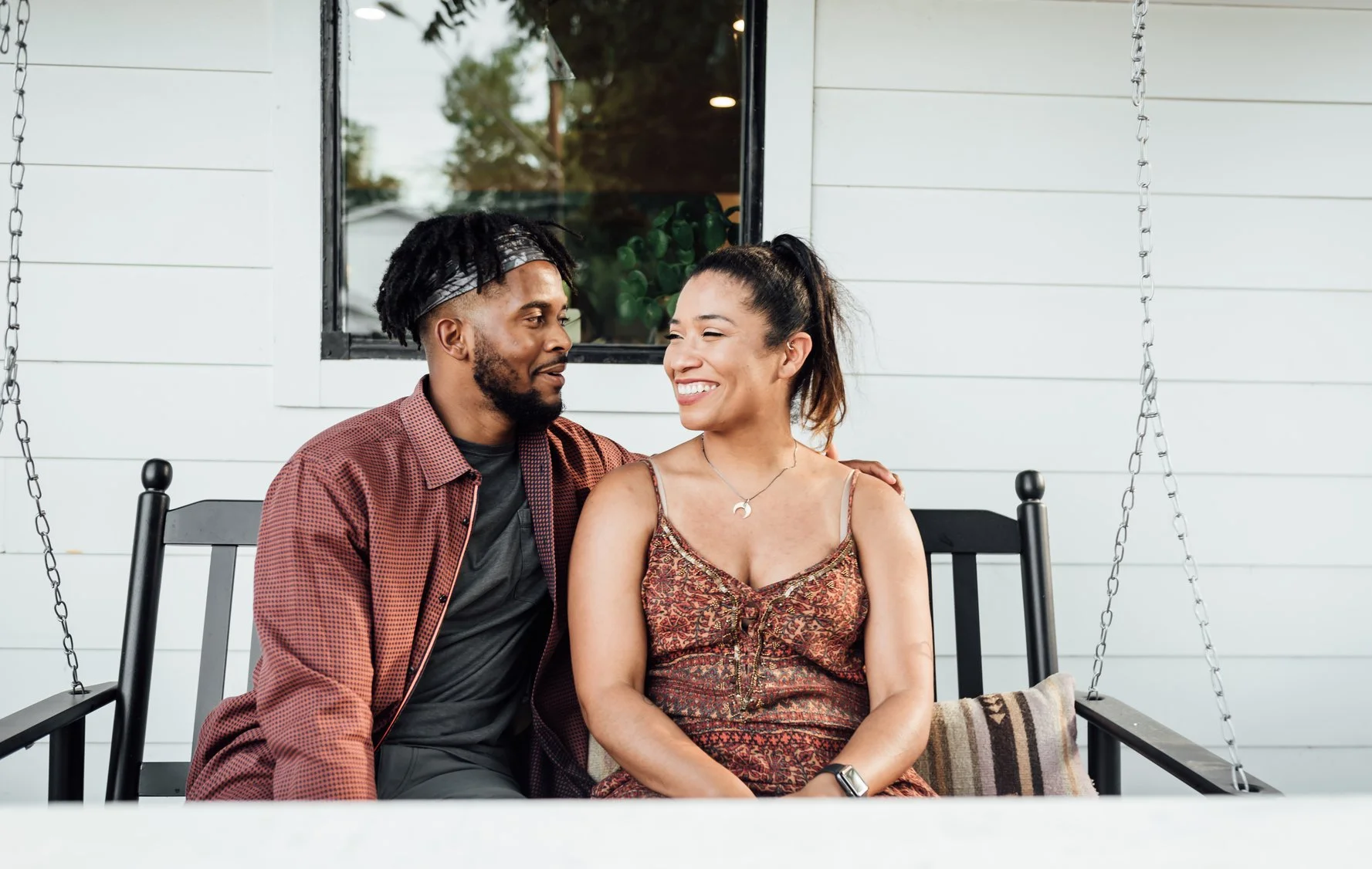 A couple sitting on a porch swing, smiling at each other. The man has dark hair styled in dreadlocks with a headband, wearing a red checkered shirt. The woman has dark hair in a ponytail, wearing a patterned dress and necklace. EFT Couples Therapy