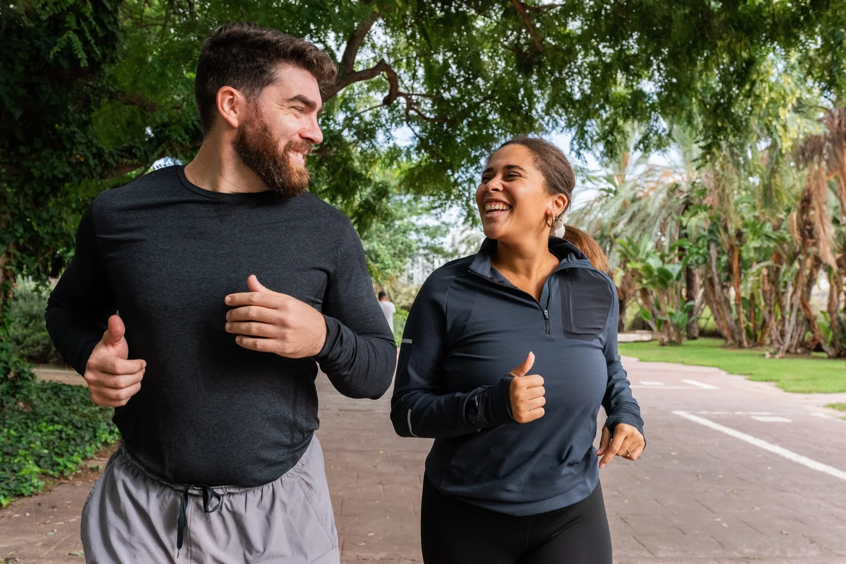 A man and a woman jogging outdoors on a park path, smiling and looking at each other, surrounded by green trees. EFT Couples Therapy in Royal Oak, Michigan and online statewide