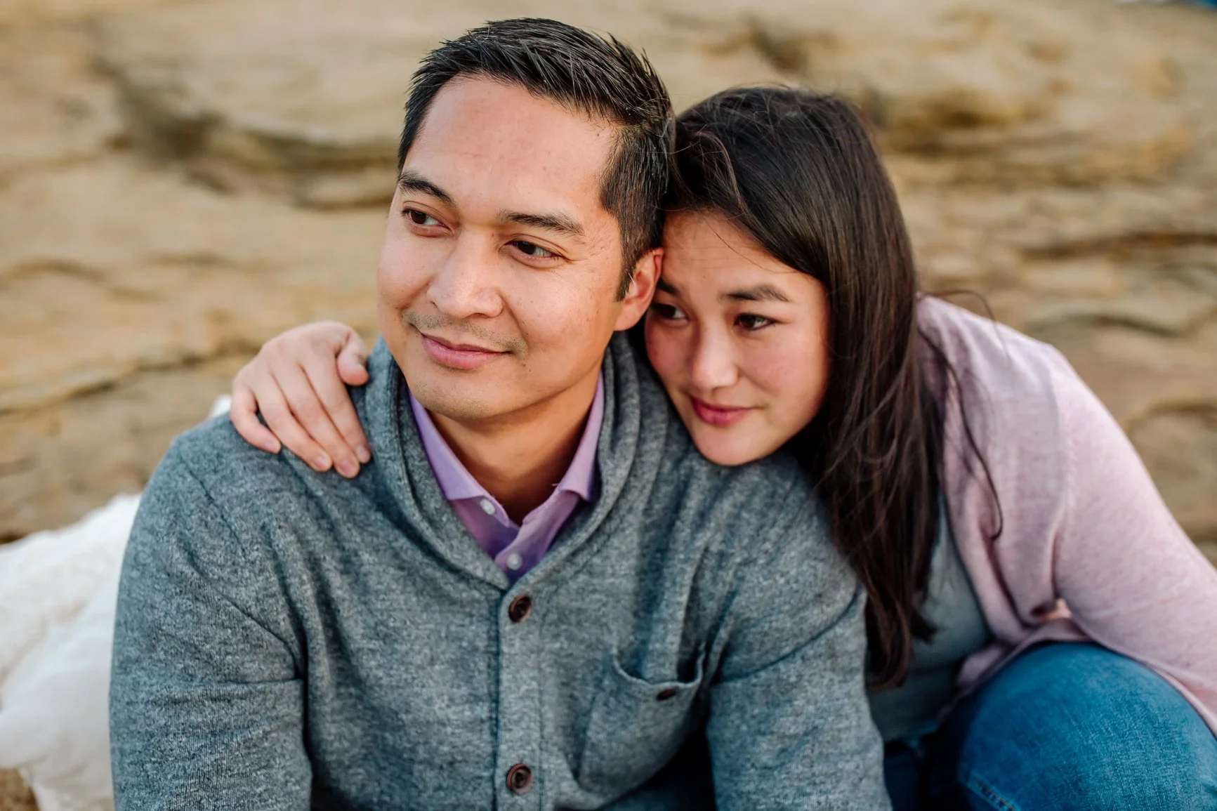A man with short dark hair and wearing a gray sweater and purple shirt sits on a rocky beach, with a woman with long dark hair and wearing a pink cardigan and blue jeans leaning on his shoulder, smiling gently. EFT Couples Therapy in Royal Oak, MI